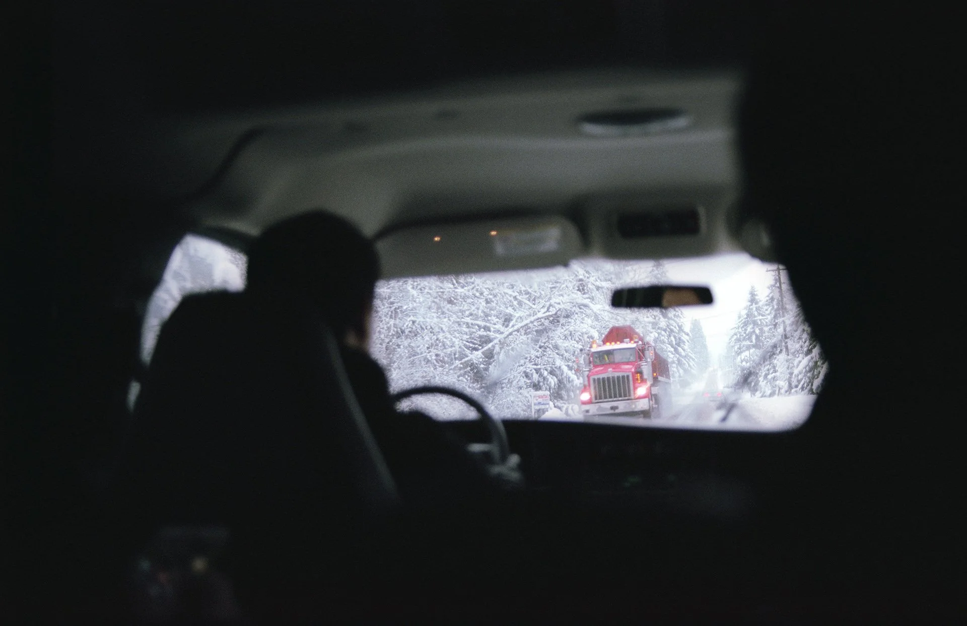 View from inside a vehicle showing a snowy winter road with a red truck in front, and snow-covered trees on either side.