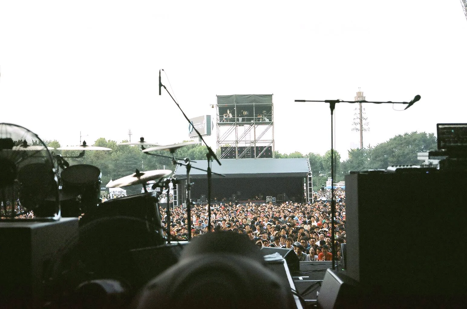 View from stage at a concert with drums and microphones, overlooking a large crowd outdoors, with a stage and lighting rig in the background.