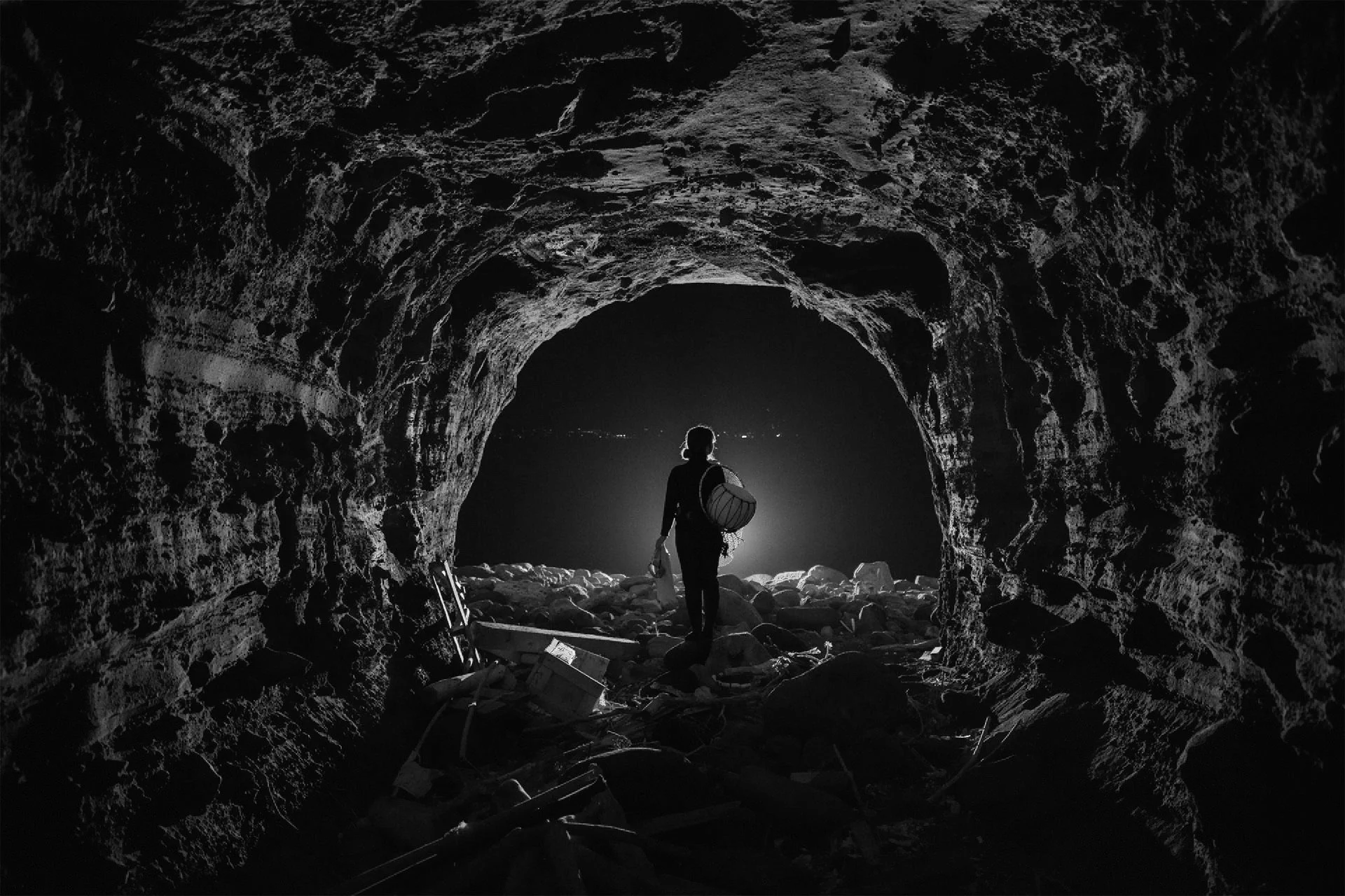 A person standing at the entrance of a dark cave, silhouetted against light coming from outside, carrying a backpack and walking on rocky ground.