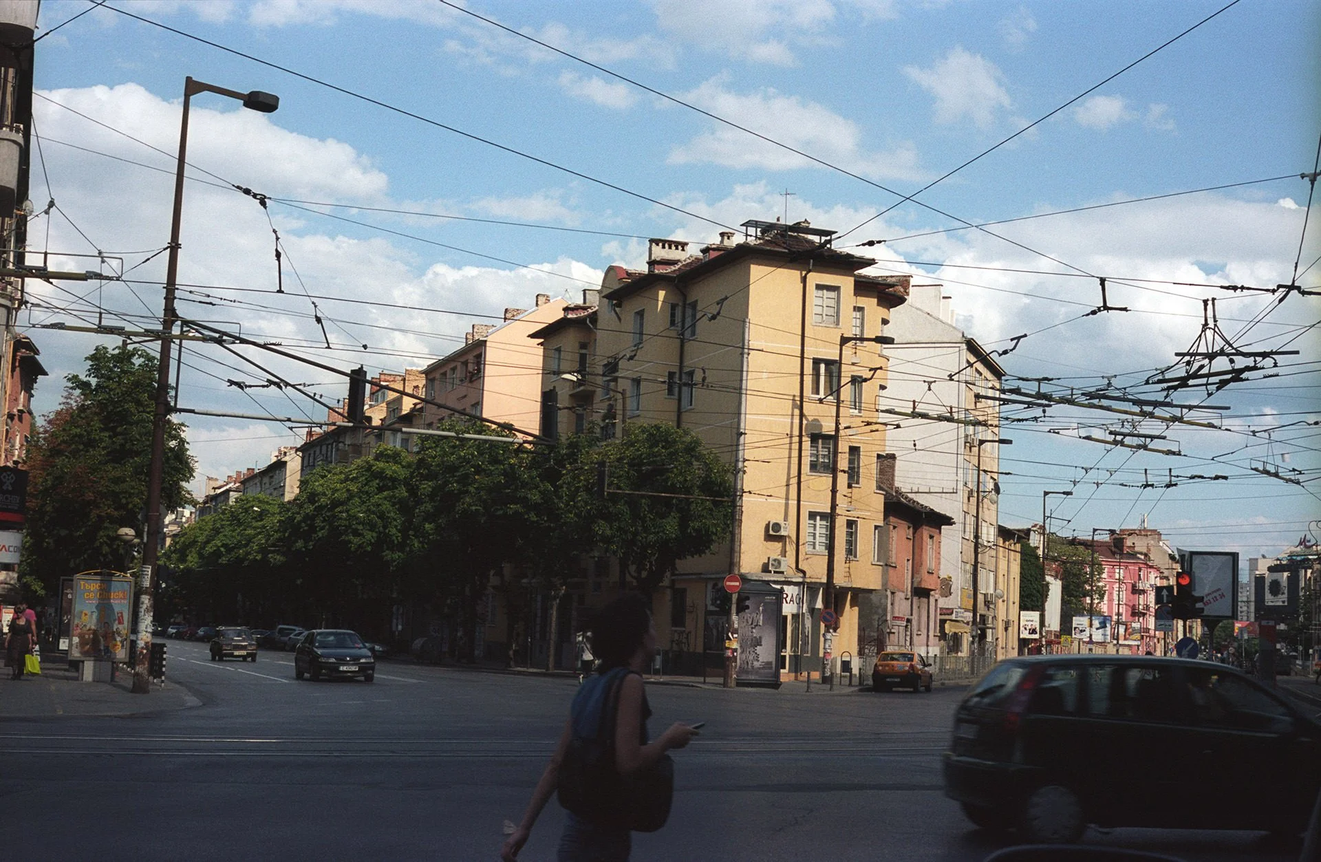 City street scene with cars, pedestrians, multi-story buildings, streetlights, and overhead tram wires under a partly cloudy sky.