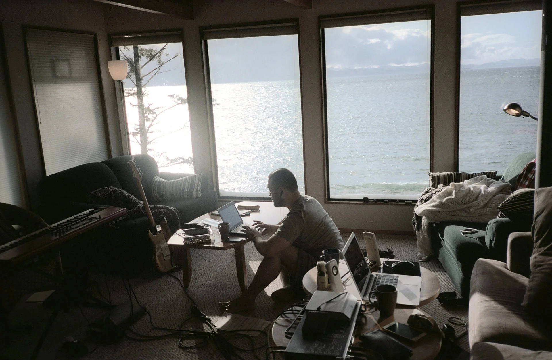 Living room with large windows overlooking water, a person working on a laptop at a coffee table surrounded by musical instruments, electronic equipment, and clutter.