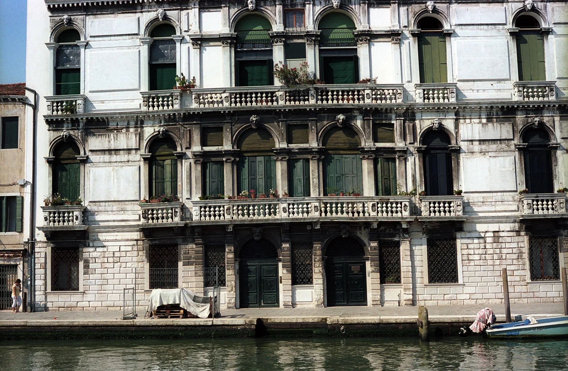 Historic building with ornate architectural details along a canal, featuring arched windows with green shutters and stone balconies, with a small boat docked nearby.