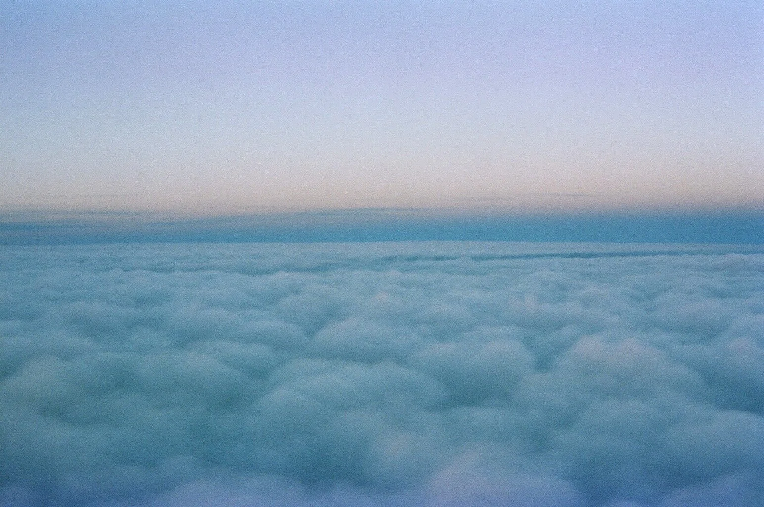 A view from above the clouds at sunset or sunrise, with soft pastel colors and a horizon line dividing the sky and cloud layer.