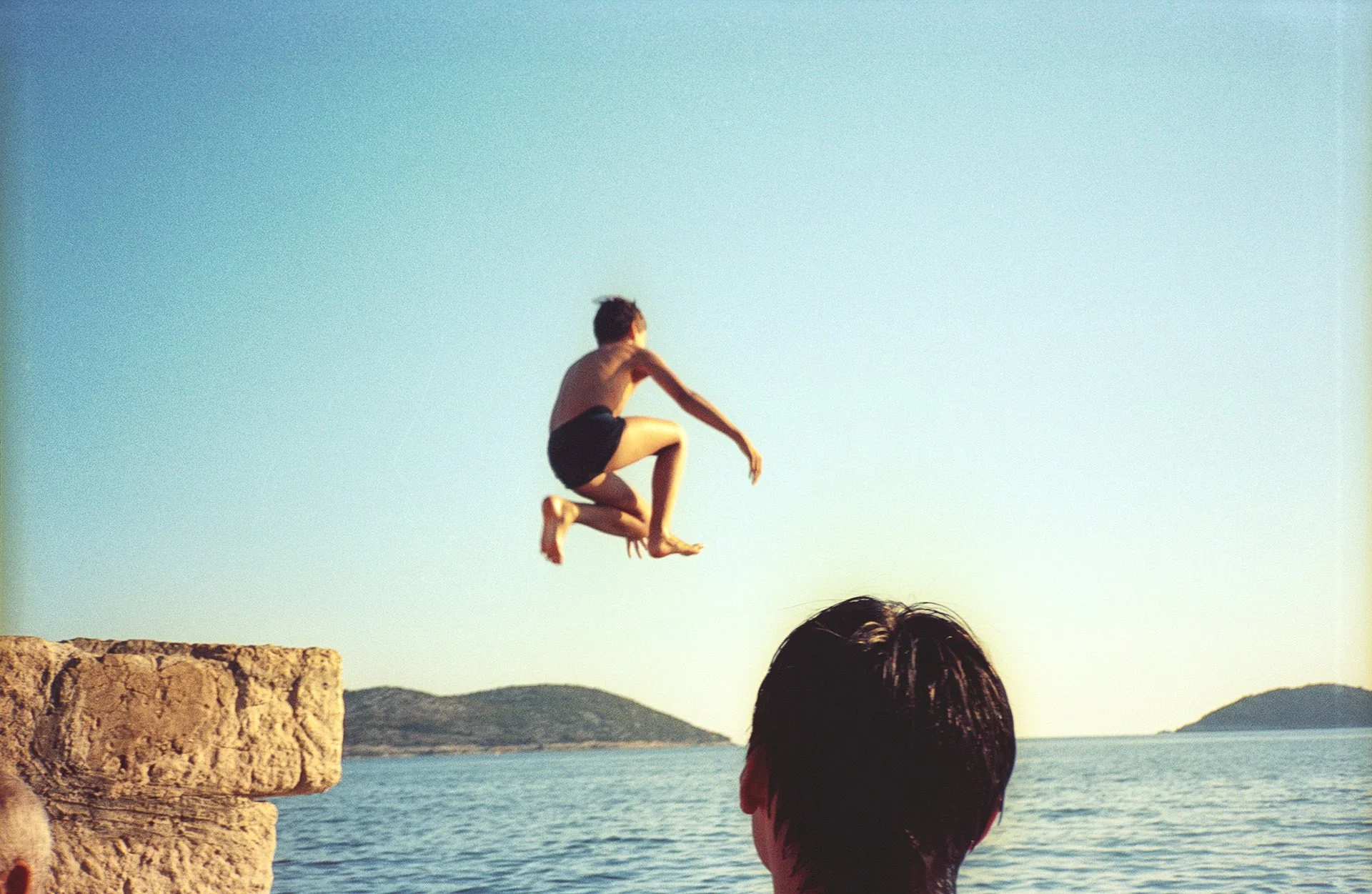 A person jumping off a stone ledge into the water with mountains in the background, viewed from behind by a person with dark hair.