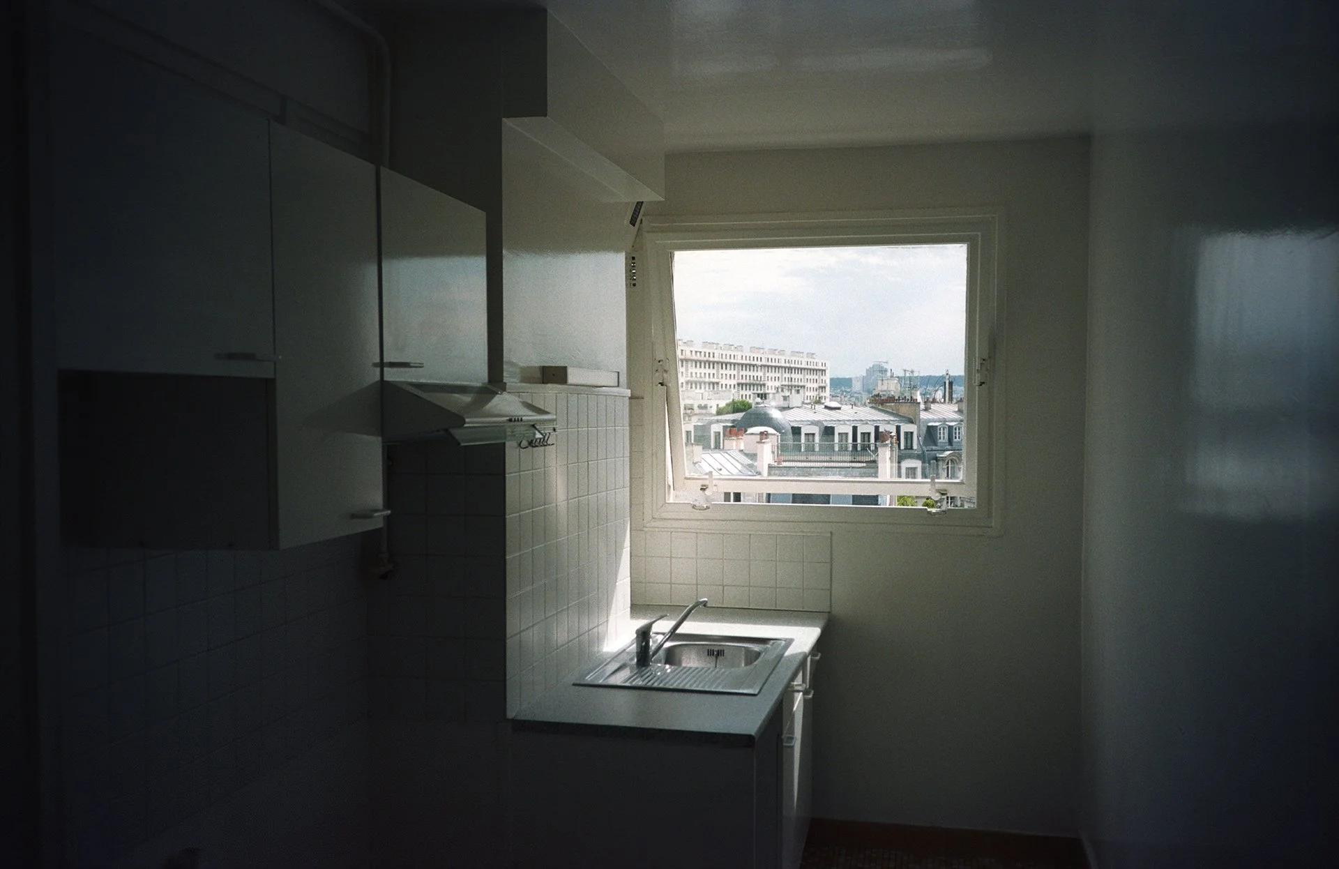 Small kitchen with a window overlooking city buildings, white cabinets, a stove with an exhaust hood, and a sink under the window.