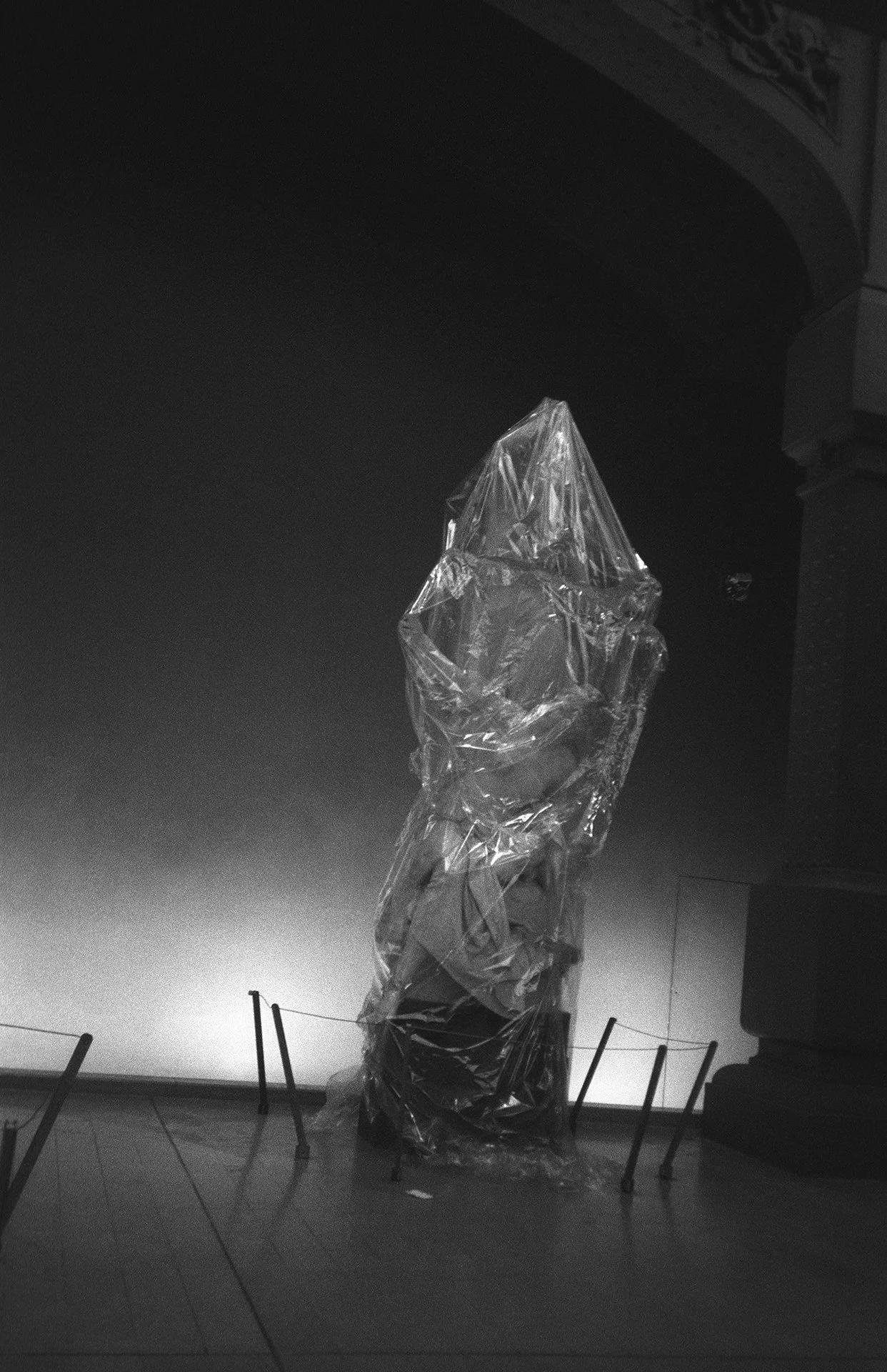 A sculpture wrapped in plastic and placed on a stand in a gallery with chairs in front. The sculpture appears to be a head or a figure.