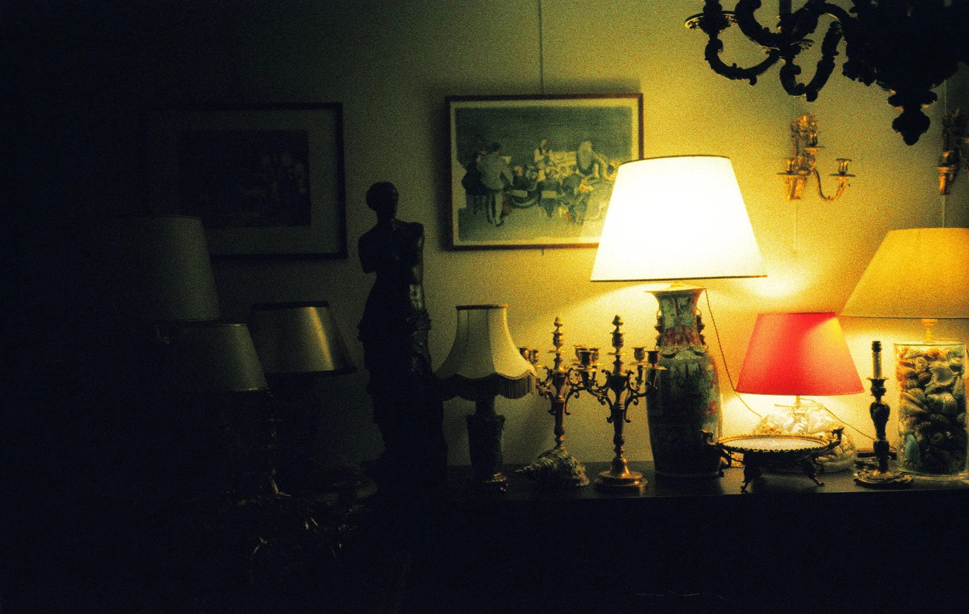 Dimly lit room with warm-colored lamps, framed artwork on the wall, a dark lamp with a floral design, a sculpture of a woman, and various decorative items on a table.