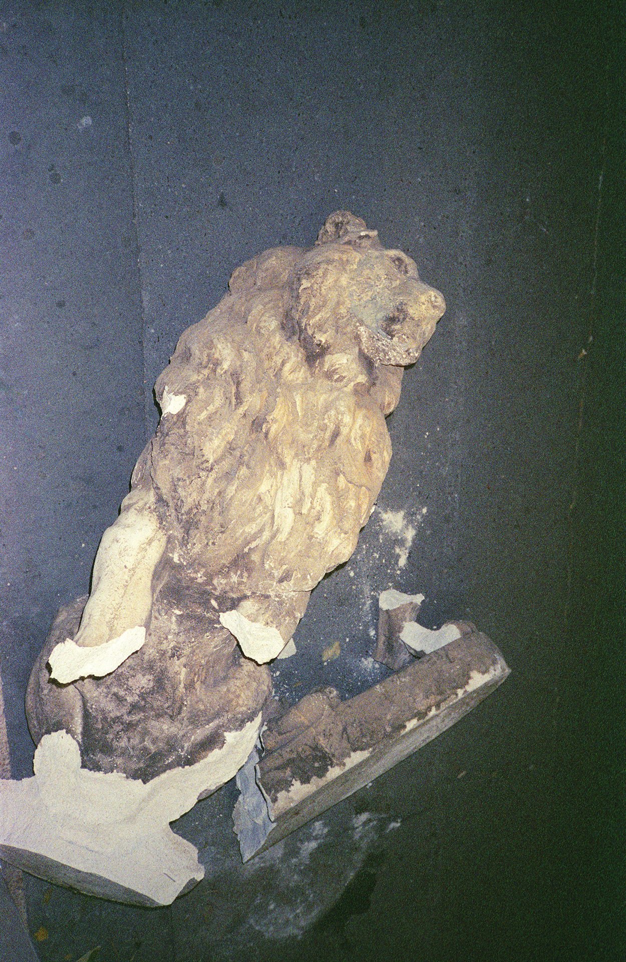 A weathered lion sculpture made of stone, sitting on a rectangular base against a dark wall background.