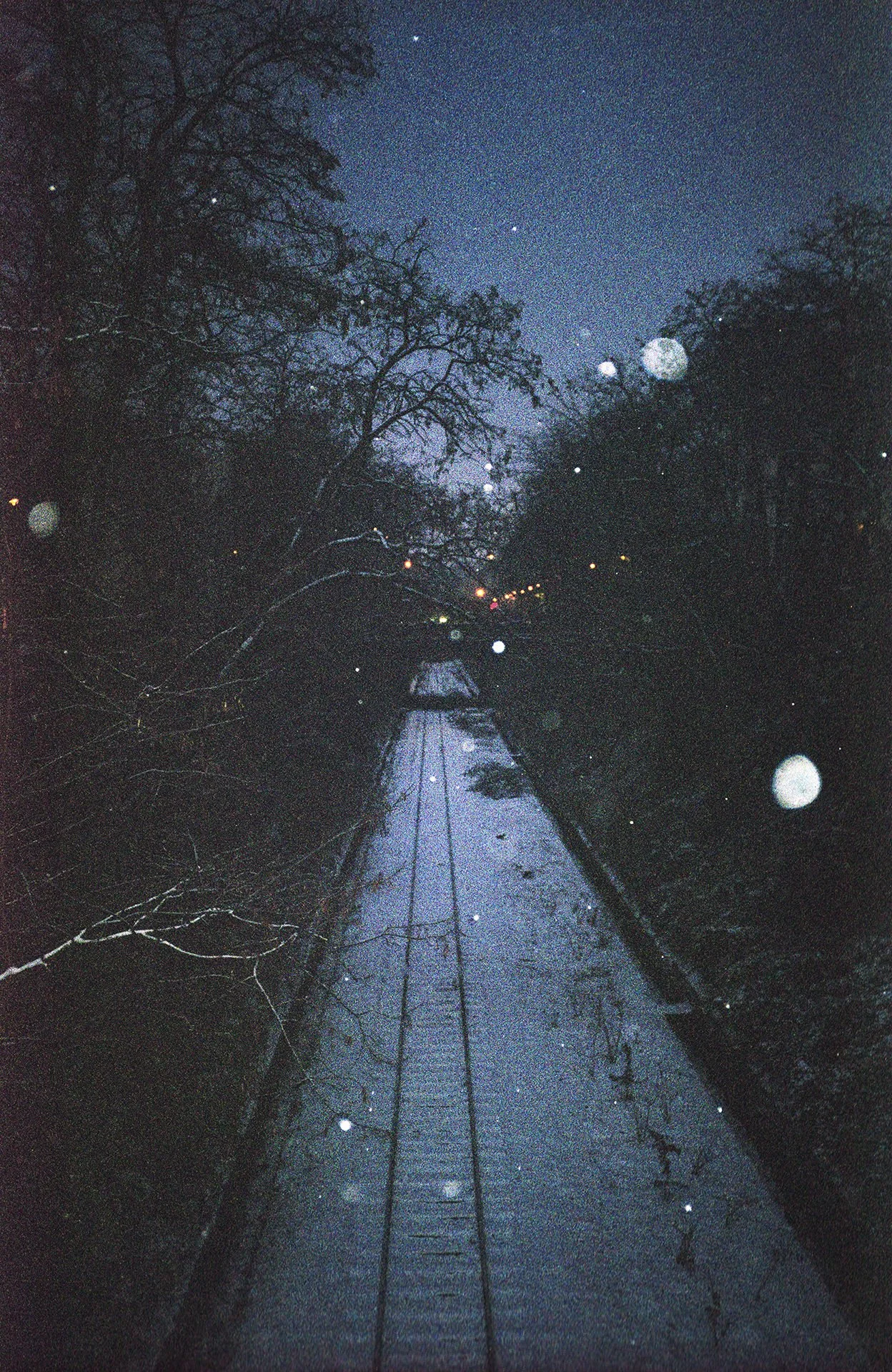 A night-time view of a narrow canal with overhanging trees and railway tracks in the middle, illuminated by distant streetlights and stars in the sky, with visible lens flare.