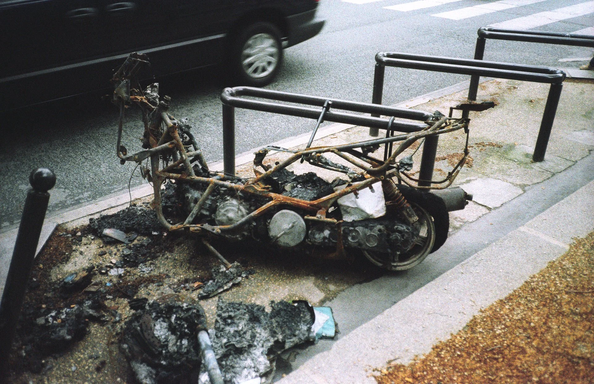 Damaged motorcycle with burnt frame and exposed engine, sitting on the sidewalk next to a street with a black car in the background.