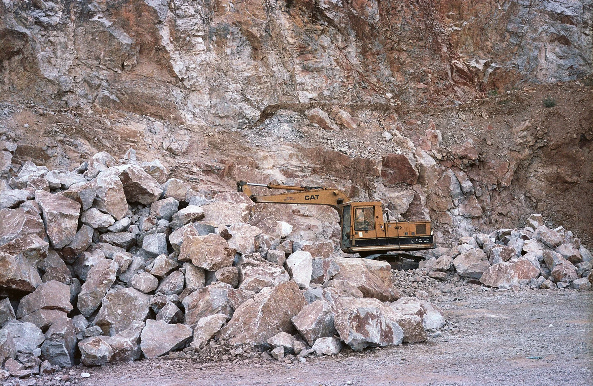 A yellow CAT excavator working at a rocky construction site with large rocks and a dirt cliff in the background.