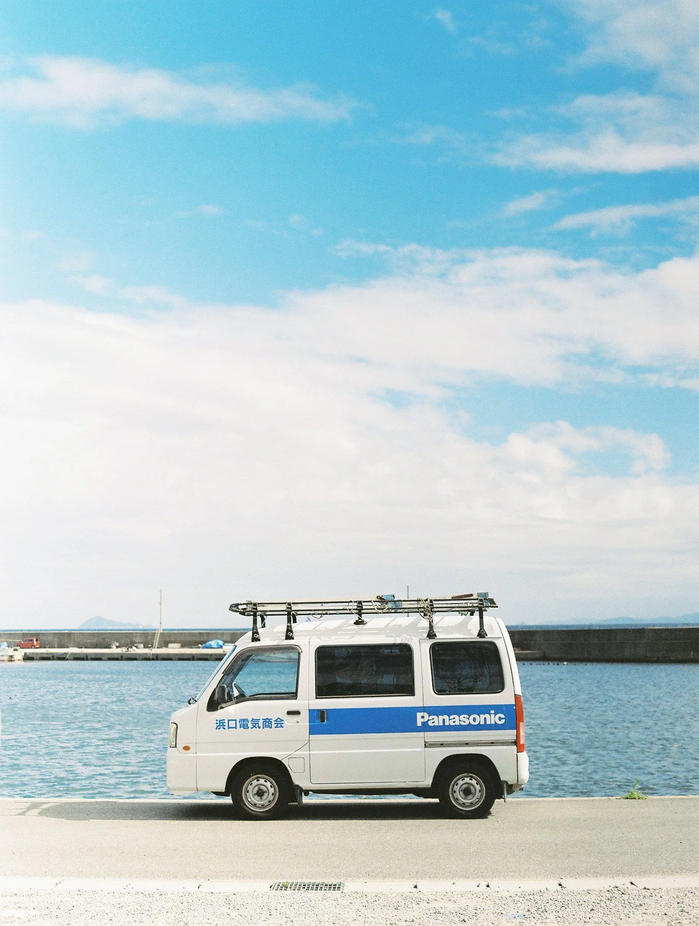 White van with Japanese writing and Panasonic logo parked near water under a partly cloudy sky.