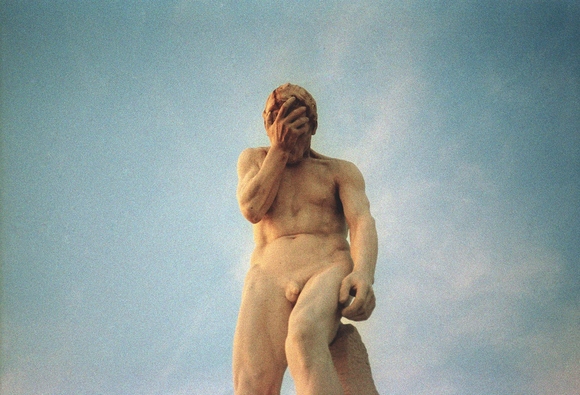 A close-up of a classical marble statue of a naked man with his hand covering his face, set against a clear blue sky.