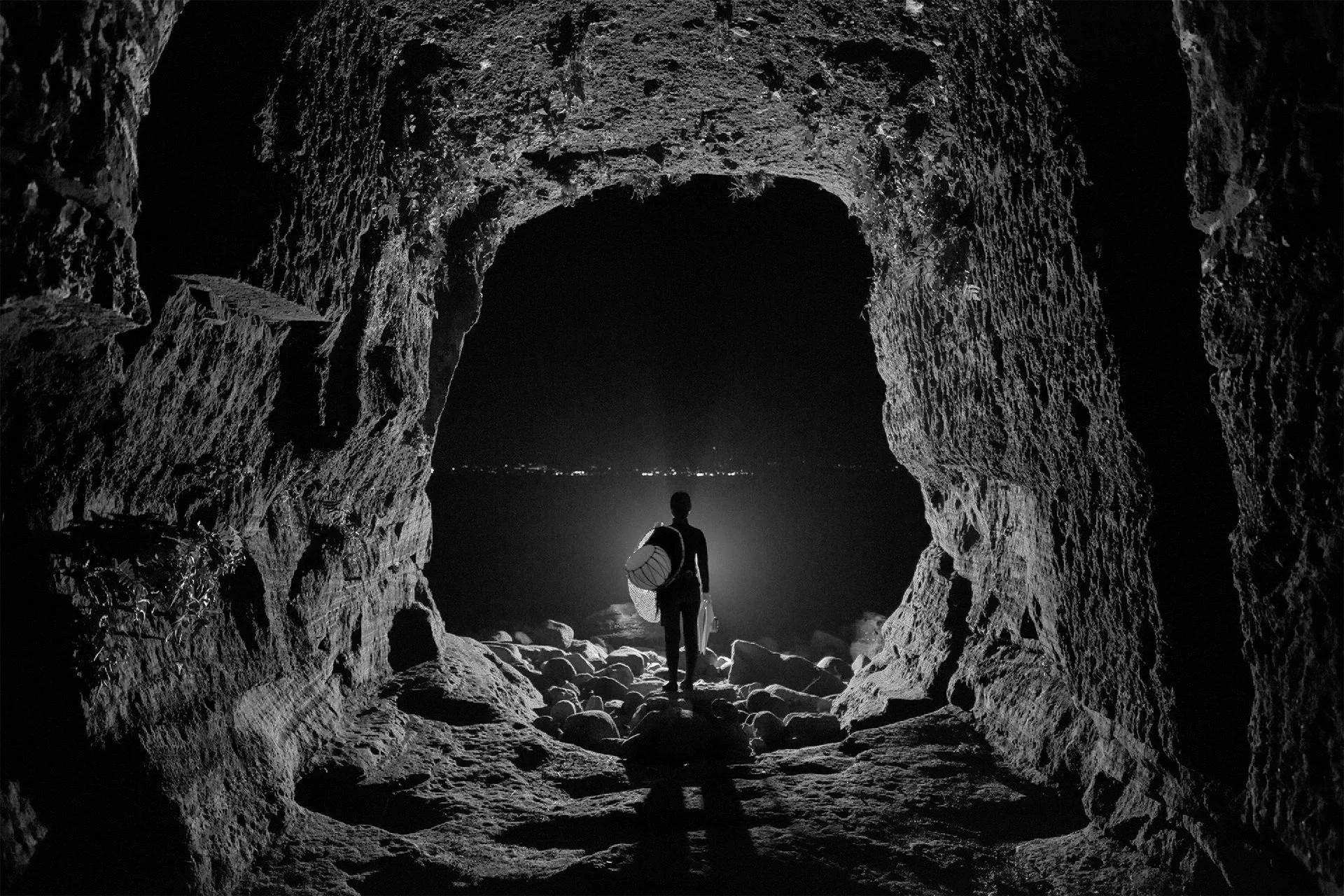 A person with a backpack and a surfboard is walking out of a large, dark cave towards the night view of a city or town illuminated in the distance.