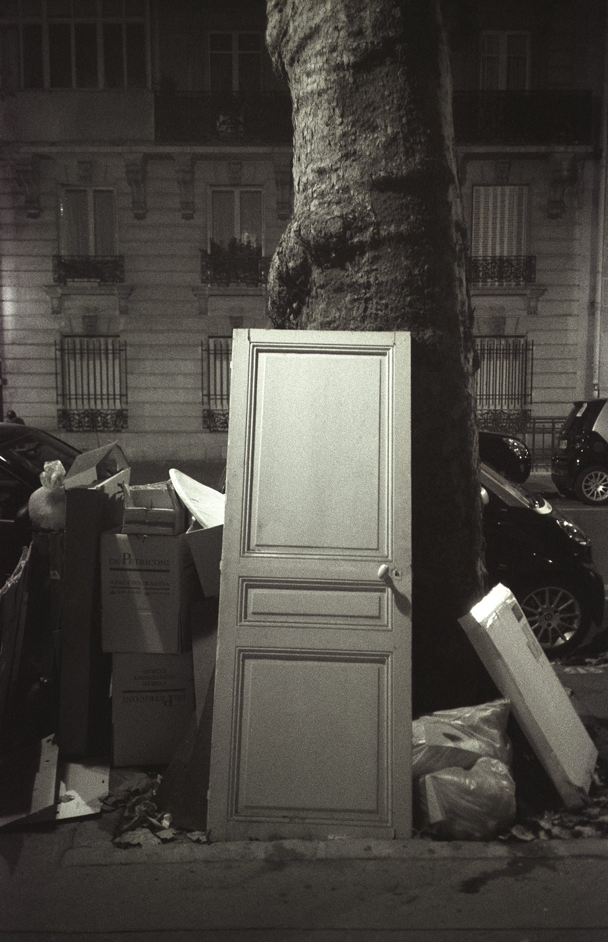 A white cabinet door leaning against a large tree trunk, surrounded by boxes, trash bags, and miscellaneous debris on a city sidewalk at night, with cars parked along the street and an apartment building in the background.