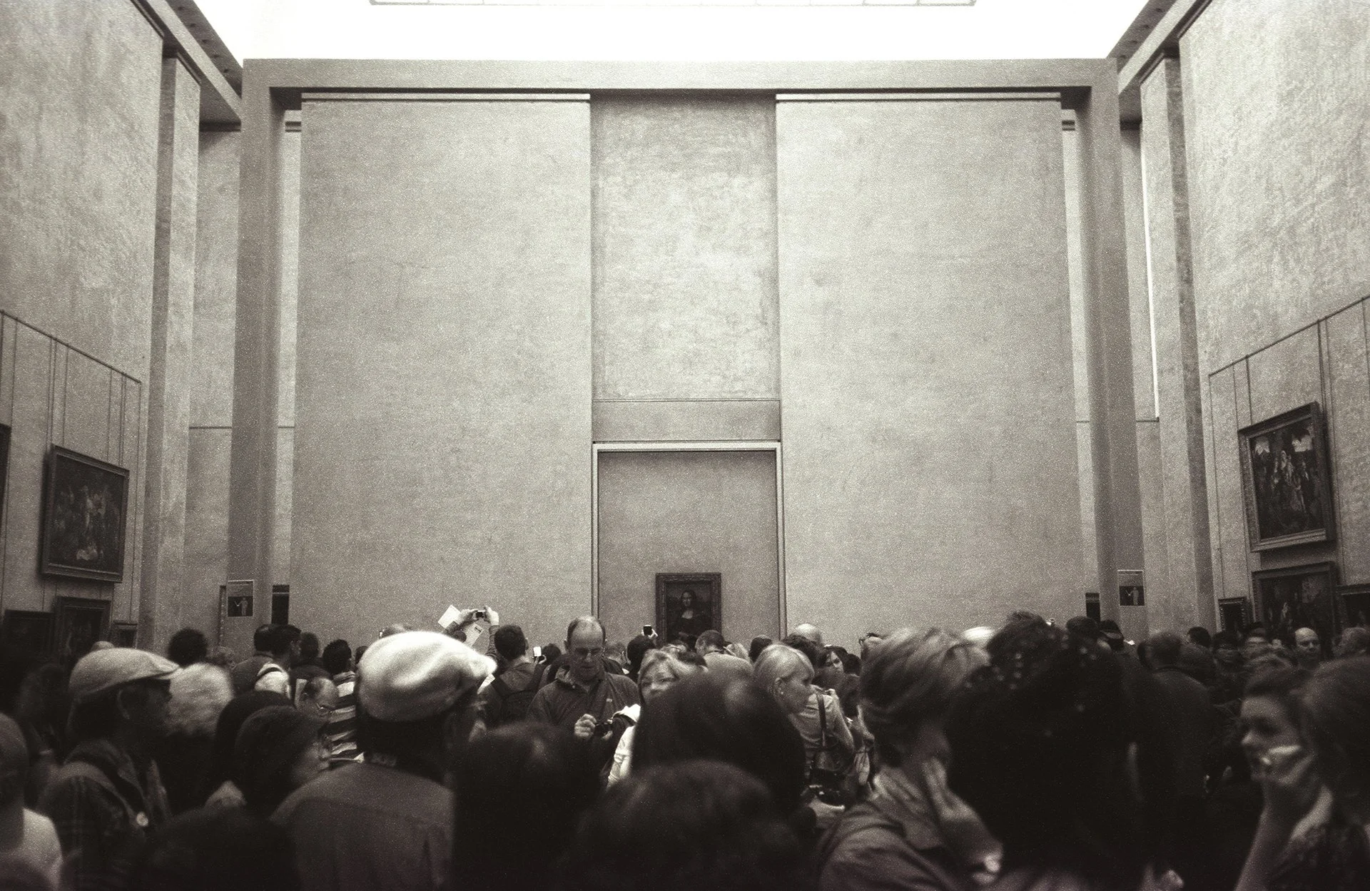 A crowded museum hall with many visitors, featuring large, framed paintings on the walls and a high ceiling with architectural details.