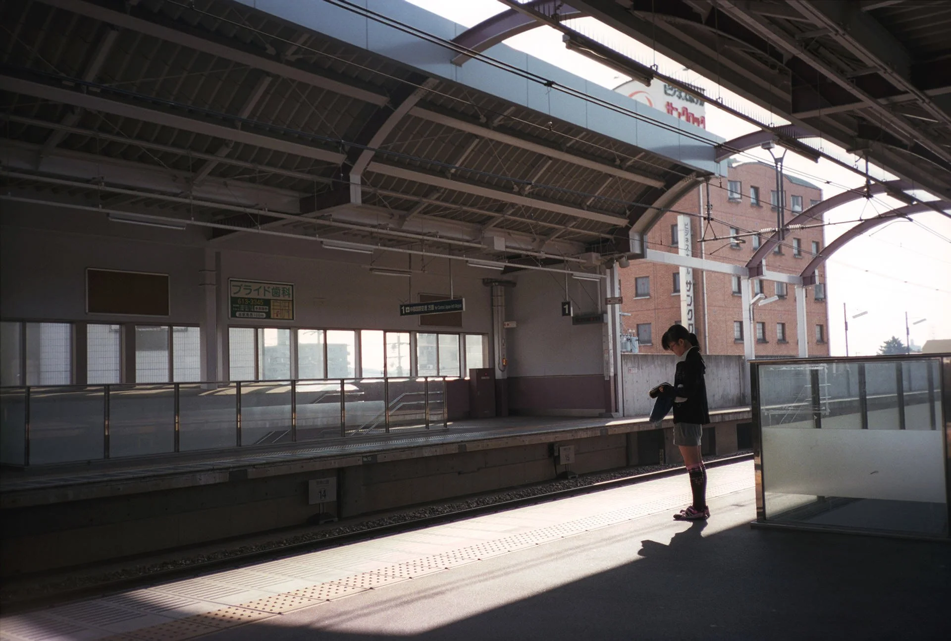 A woman wearing a black jacket, gray shorts, and pink shoes standing on a train platform, reading a book or magazine.