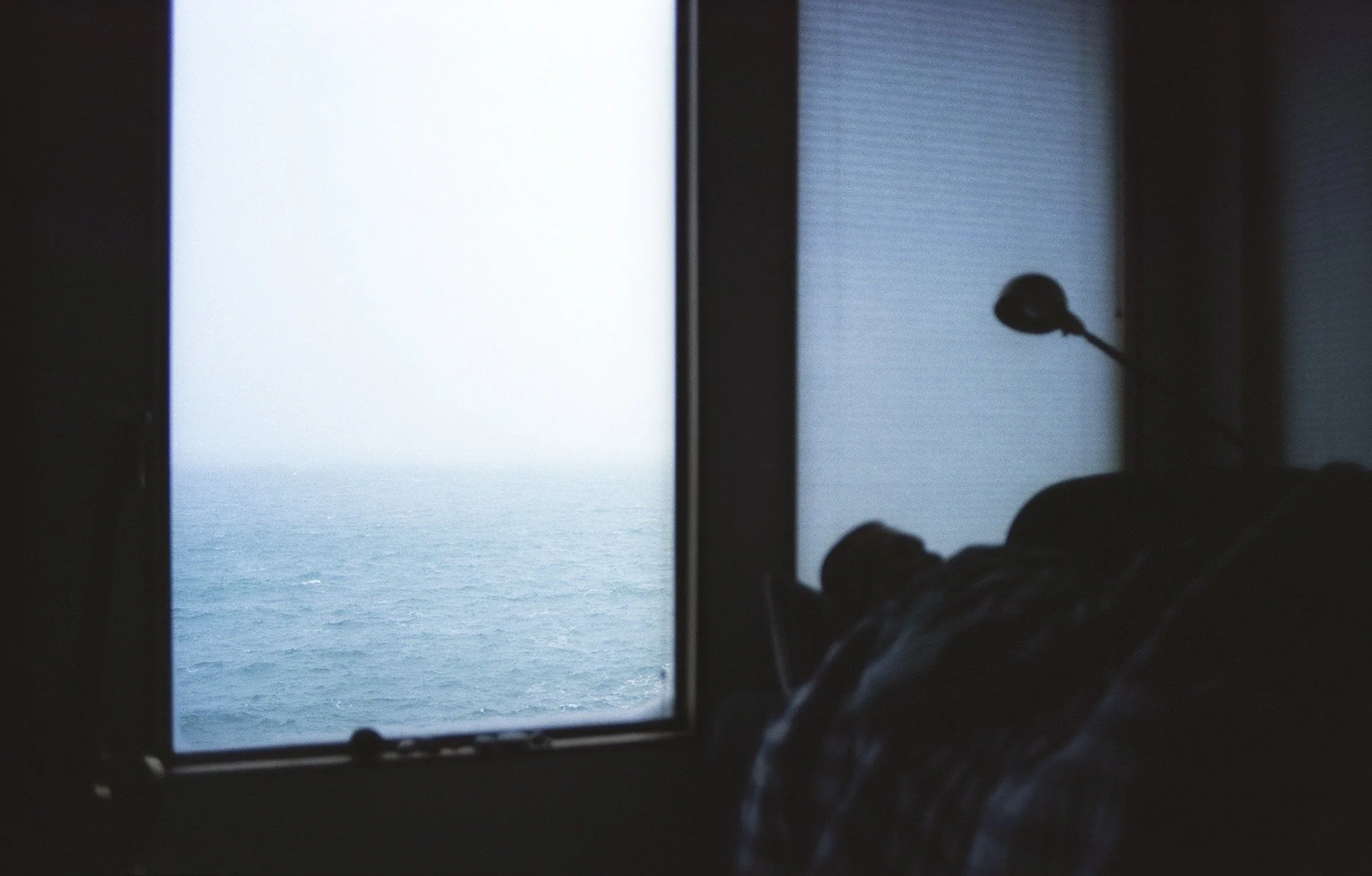 Photo taken from inside a room, looking out a window at the ocean with a partly cloudy sky, with a silhouette of a person lying down and a microphone in the foreground.