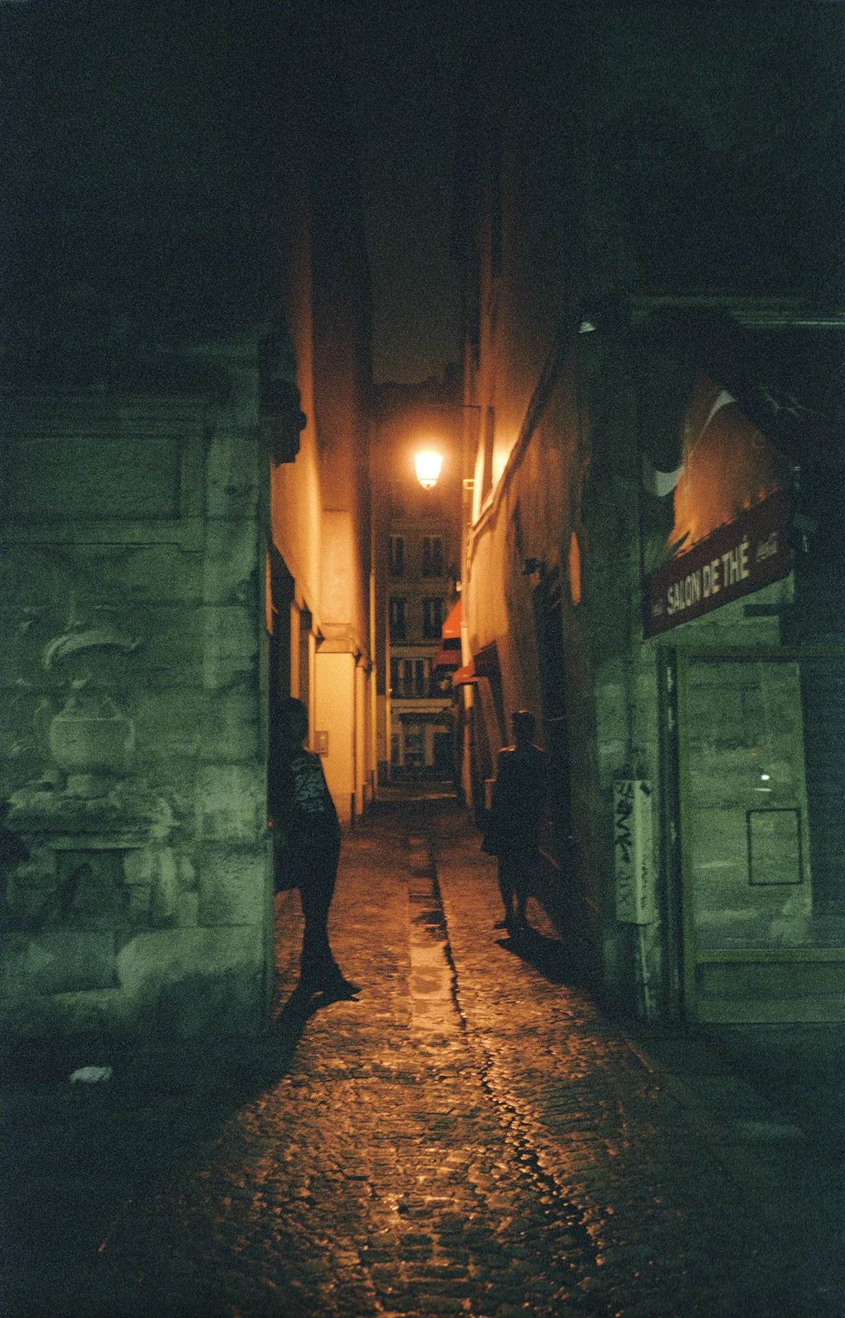 Nighttime alleyway illuminated by a street lamp, with cobblestone pavement and two people walking away from the camera.