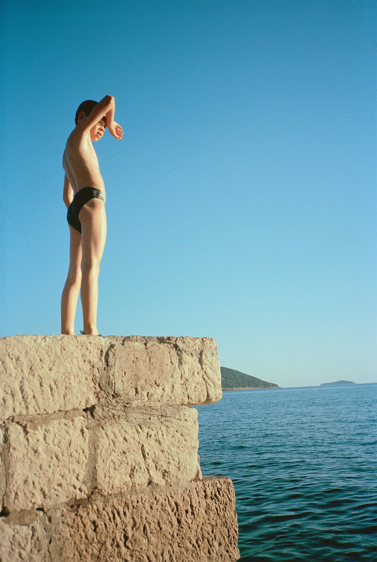 A young boy standing on a stone ledge by the sea with mountains in the background, with his eyes closed and arms raised, under a clear blue sky.