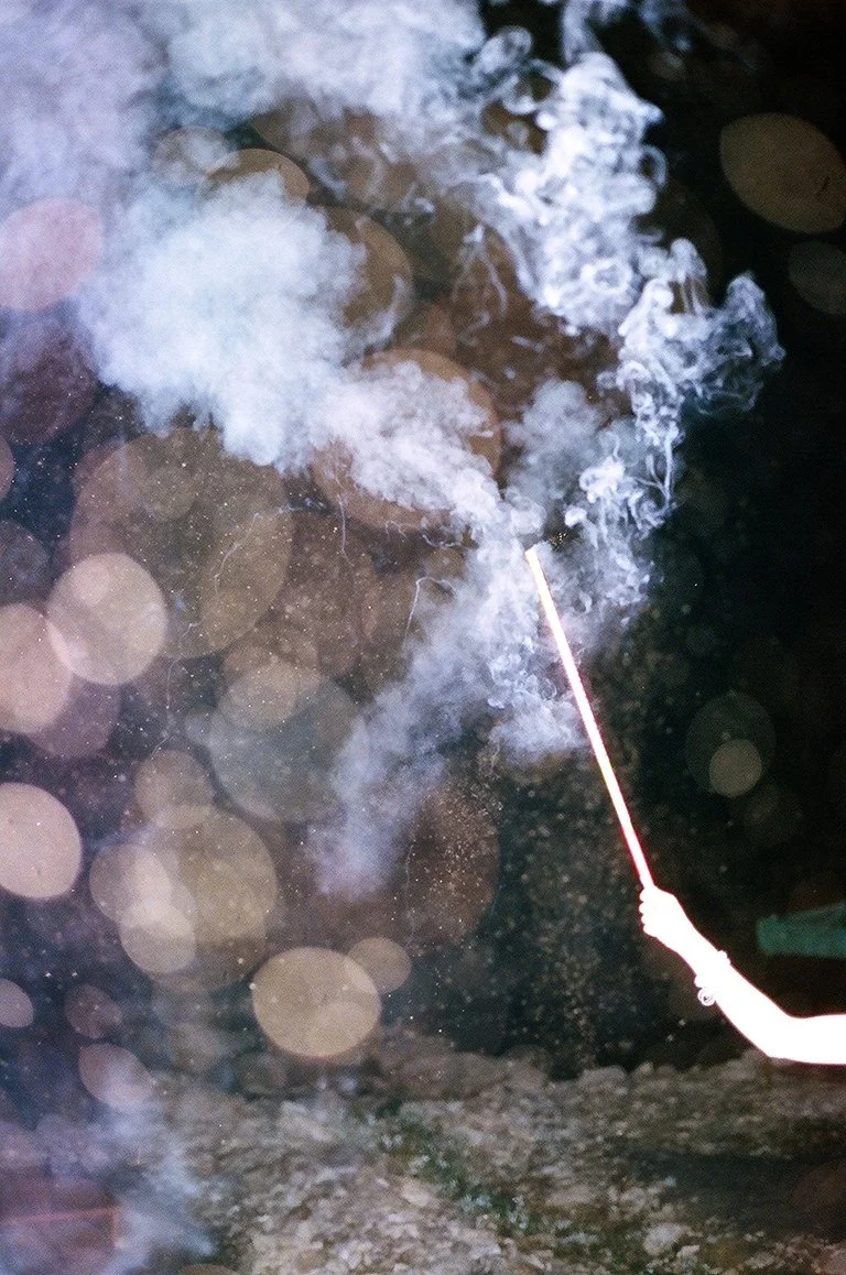 A person holding a lit sparkler over a body of water at night, with smoke rising from the sparkler and blurred circular light reflections visible in the background.