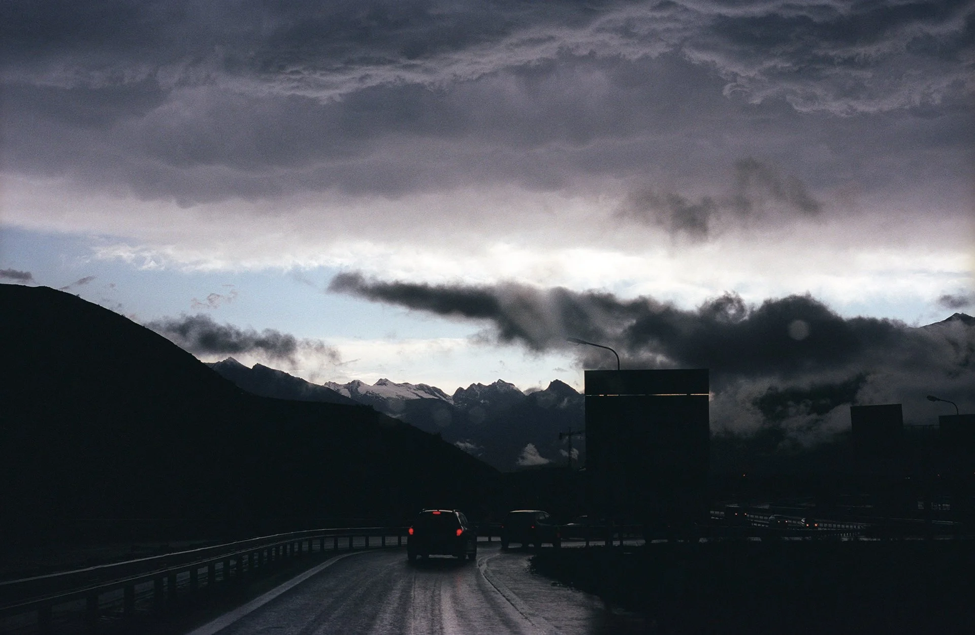Dark cloudy sky over a mountain landscape with several cars driving on a wet road and a few streetlights or road signs in the foreground.