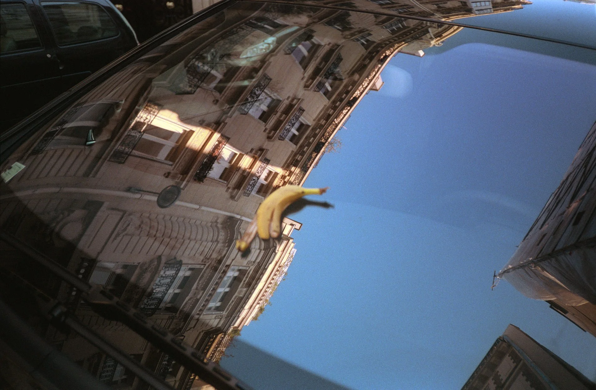Reflected image of multi-story residential buildings with balconies and windows, seen on the surface of a black vehicle outside under a clear blue sky, with a hanging yellow banana sticker on the car.