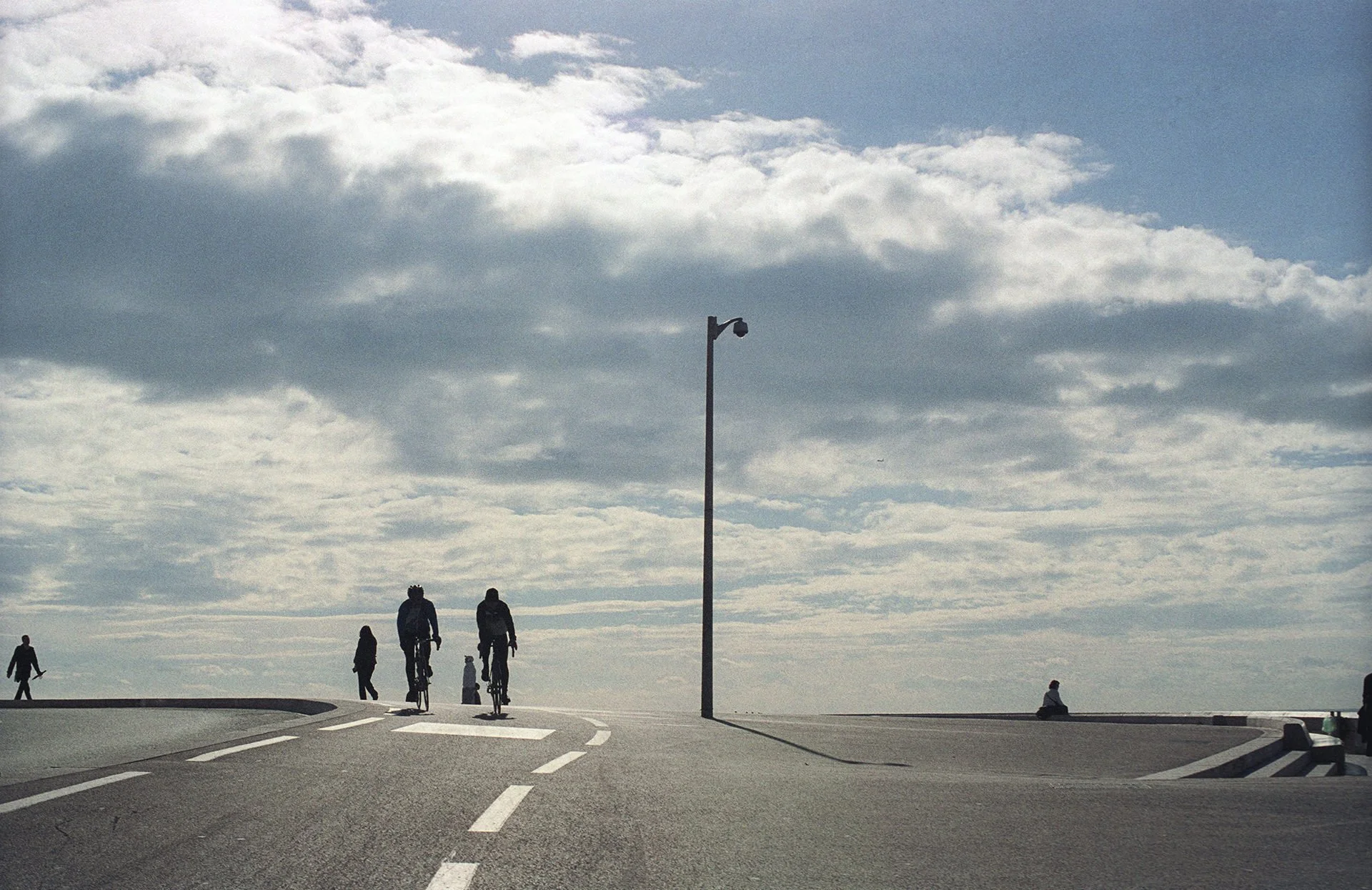People walking and cycling on a curved pathway outdoors under a cloudy sky, with a tall street lamp in the middle, some trees, and a bench on the right side.