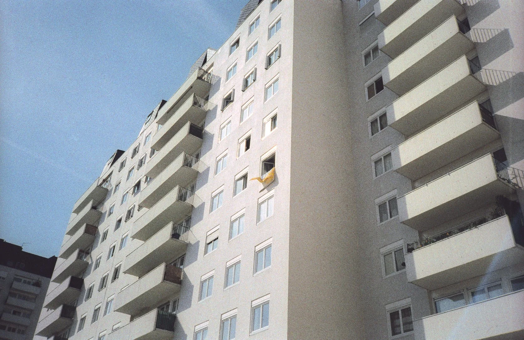 A white multi-story apartment building with rows of windows and balconies. One yellow cloth is hanging out of a window on the lower right side. The sky is clear and blue.