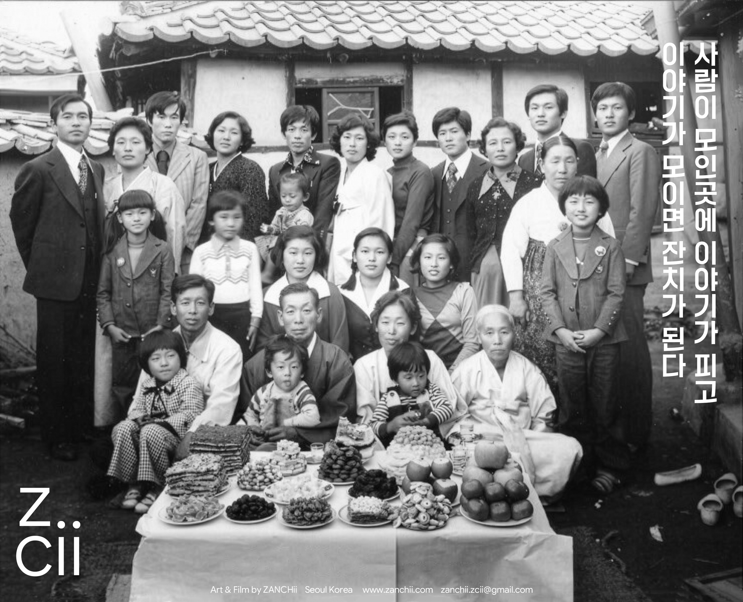 A black-and-white photograph depicting a group of Korean people, including adults and children, standing together in front of traditional Korean houses. In front of them, a table is filled with various foods and fruits, possibly set up for a celebrat