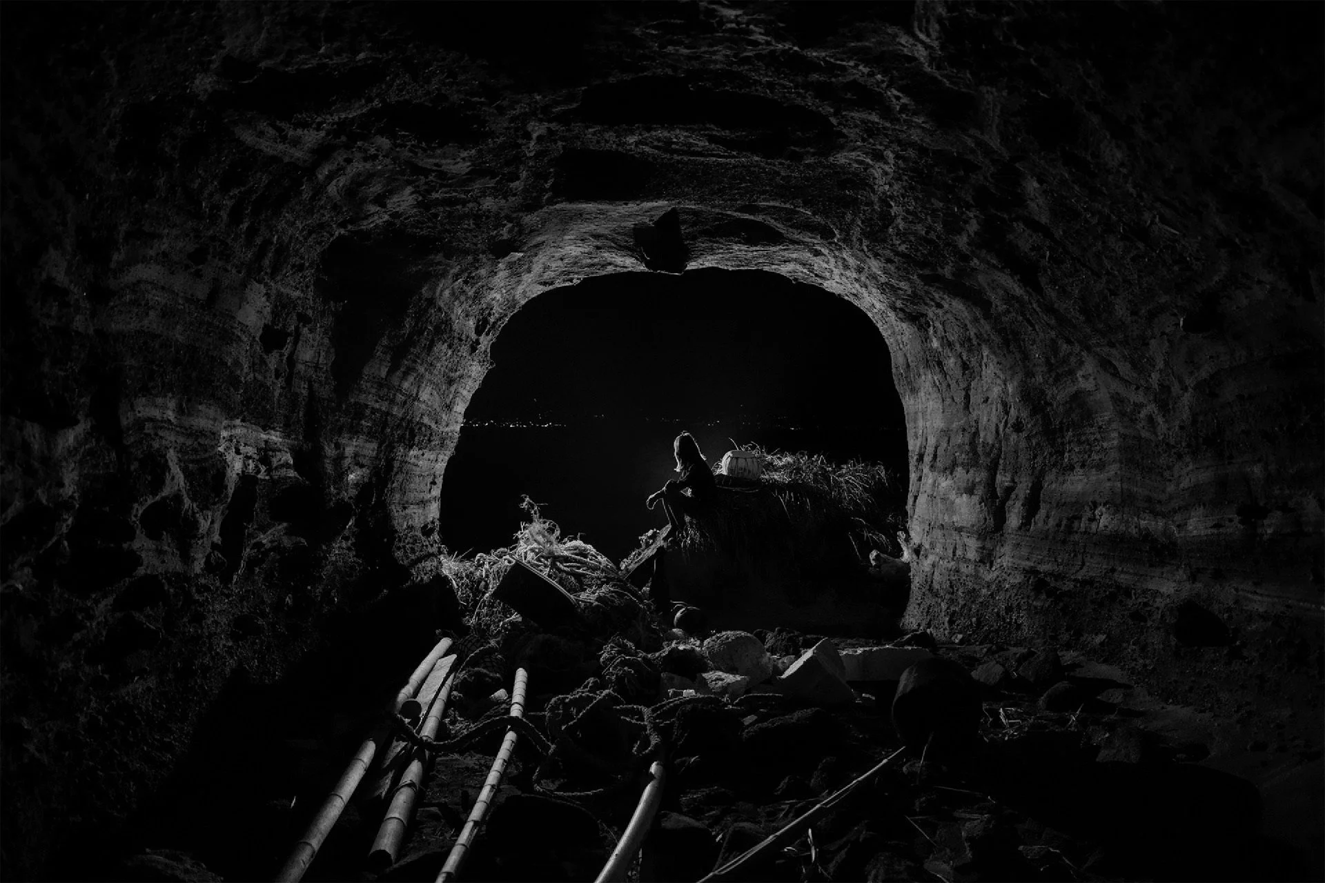 A person sitting inside a dark cave at night, with a cityscape visible in the distance through the cave opening.