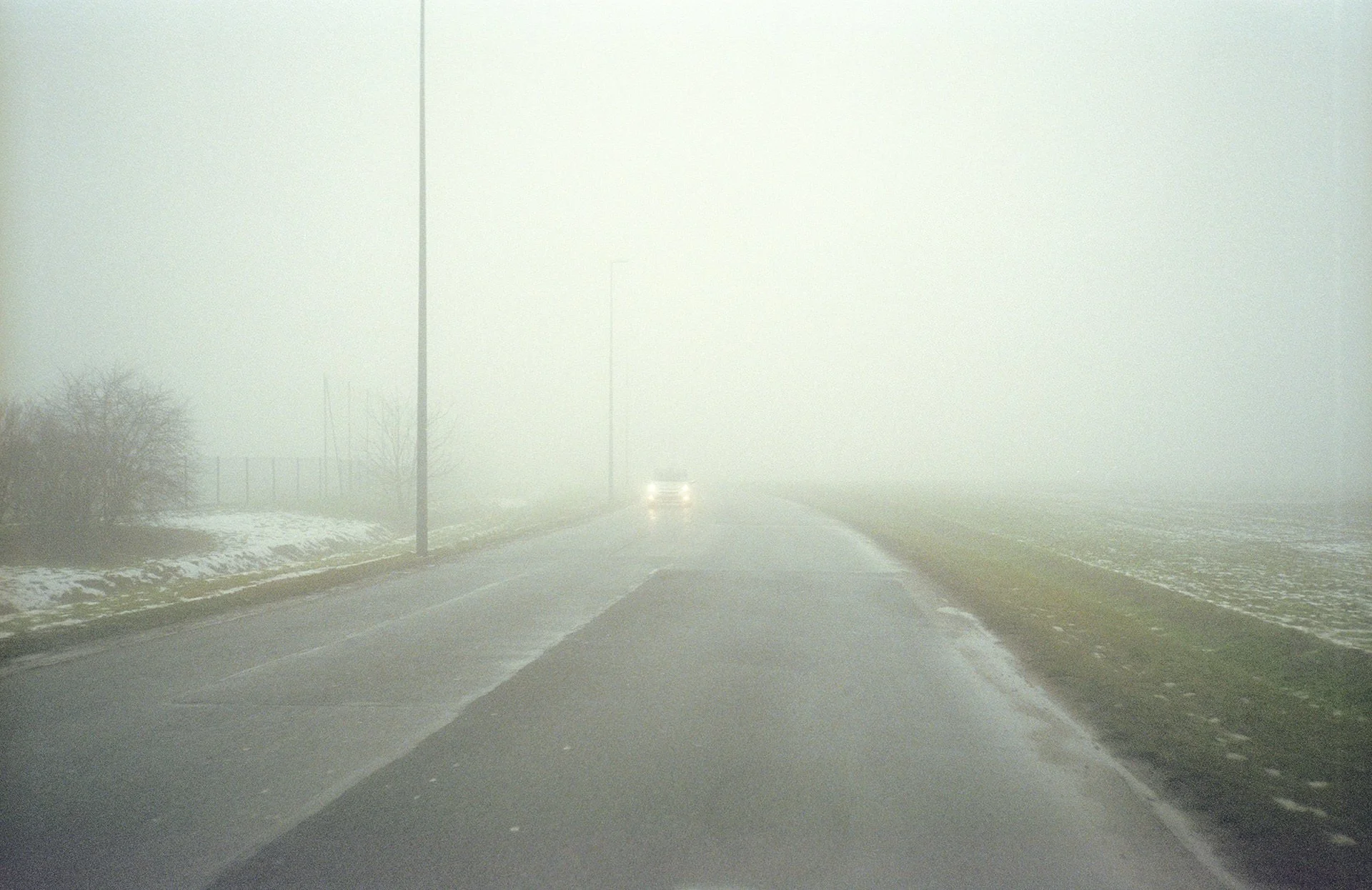 A foggy road with a car driving away in the distance, flanked by snow on the ground and utility poles lining the side.