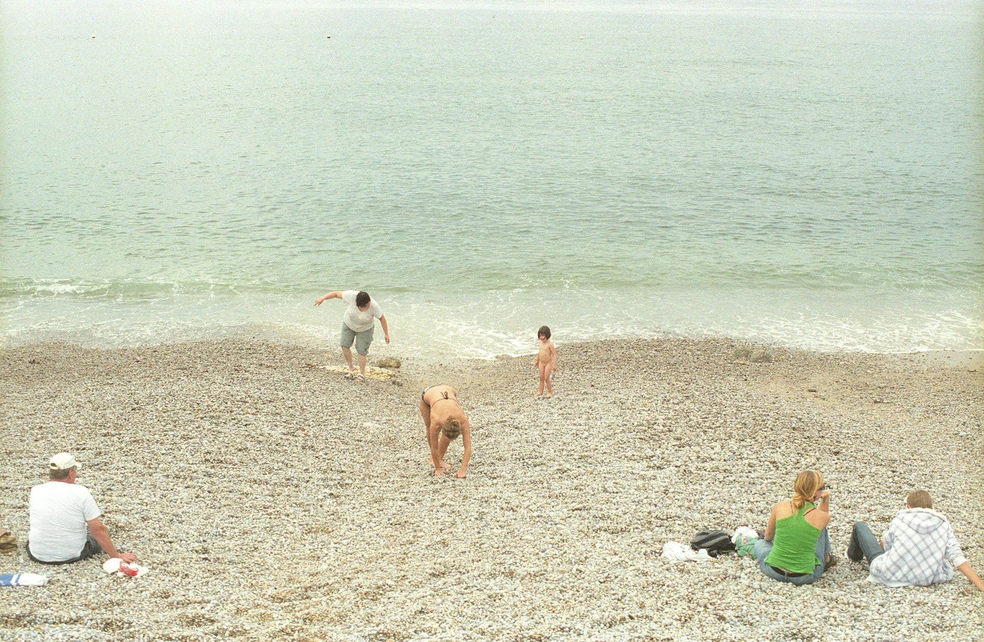 People relaxing and playing on a pebbled beach near the ocean with a few individuals wading into the water. Some are sitting on the beach, and others are standing or bending down.