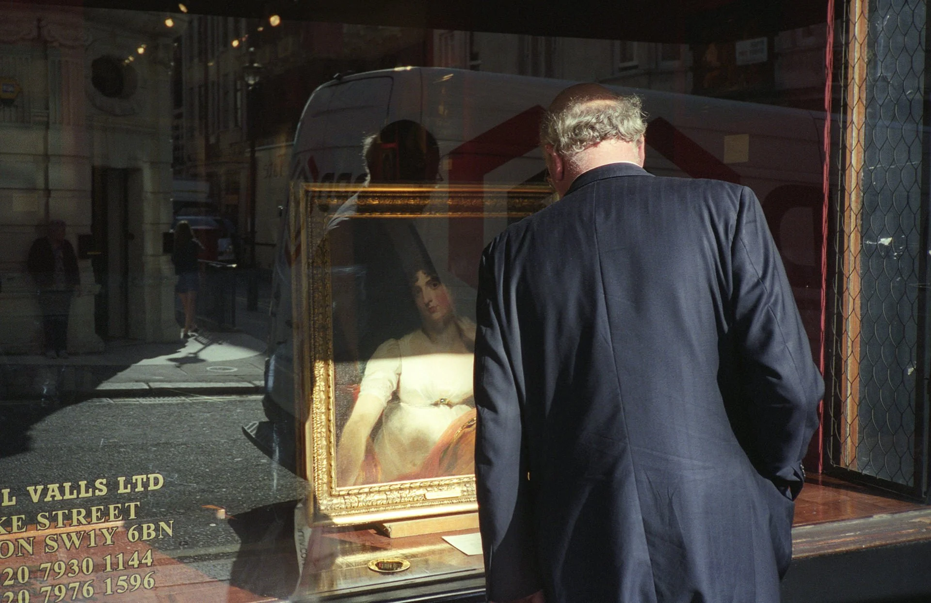 A man with curly gray hair wearing a dark suit is looking at a painting through a shop window. The painting depicts a woman with dark hair dressed in light-colored clothing. The window reflects the street scene outside, including pedestrians and buil