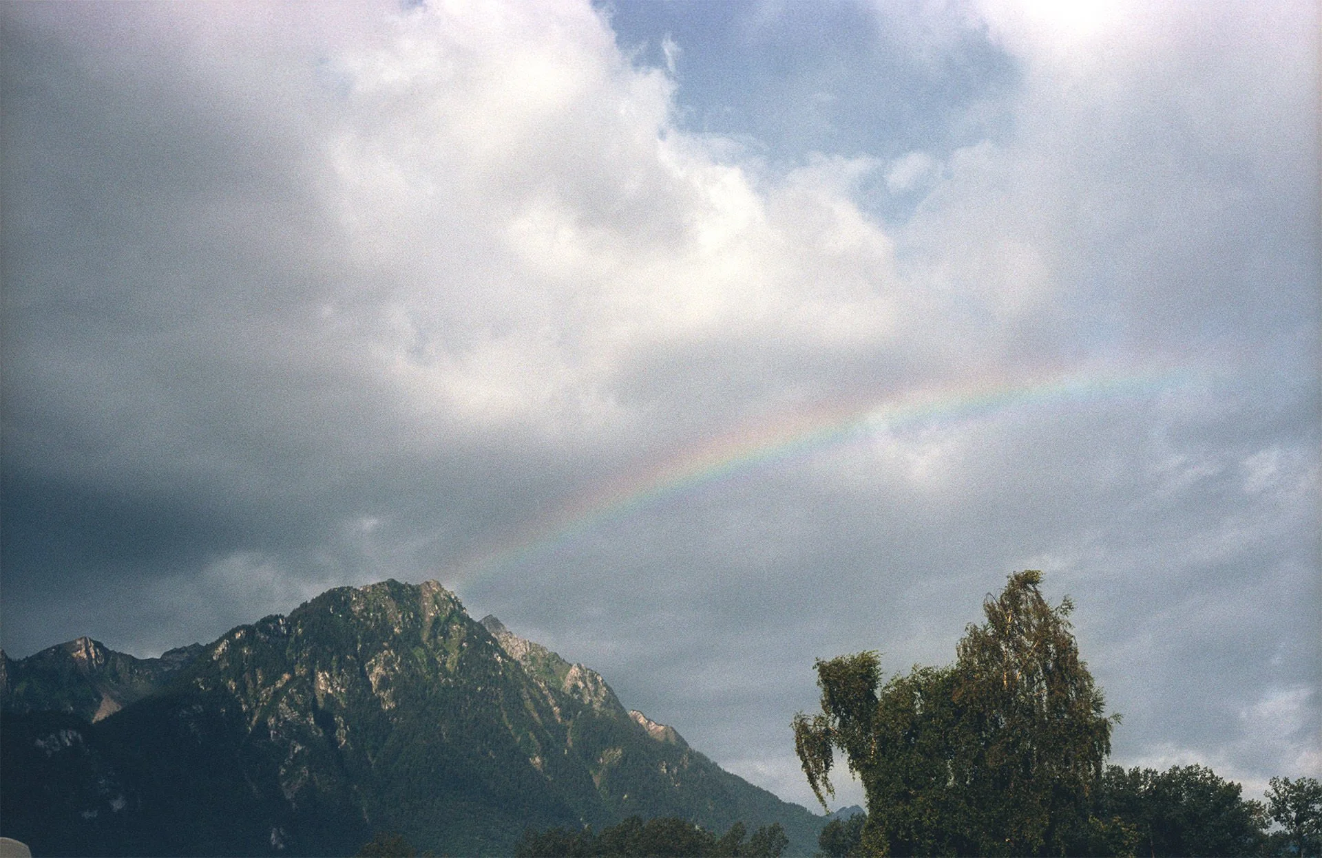 Cloudy sky over mountain range with visible rainbow and trees in the foreground.