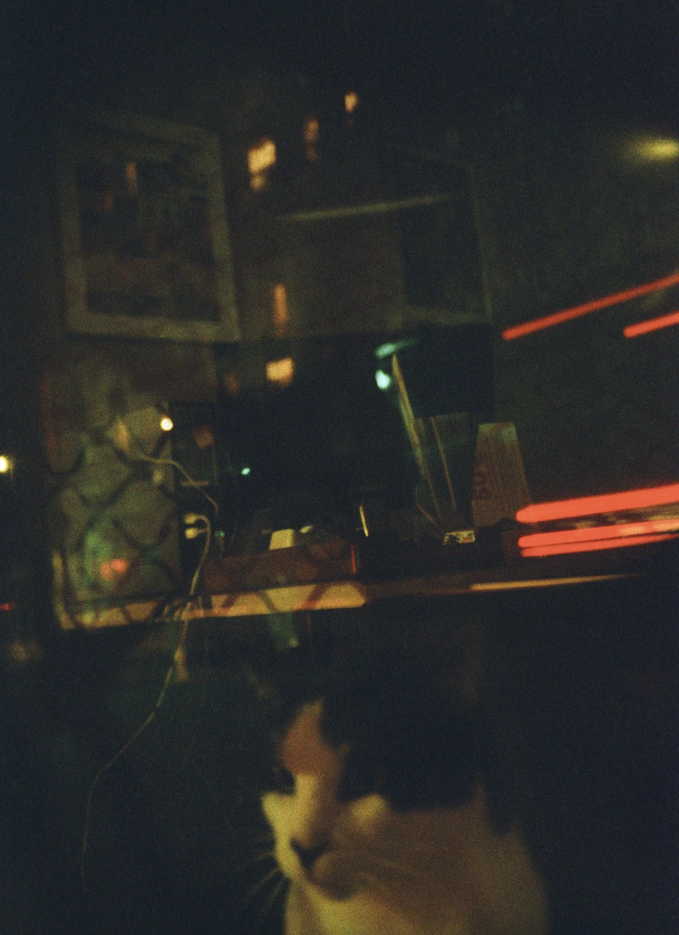 A black and white cat standing on a dark surface with a computer monitor, wires, and electronic equipment in the background.