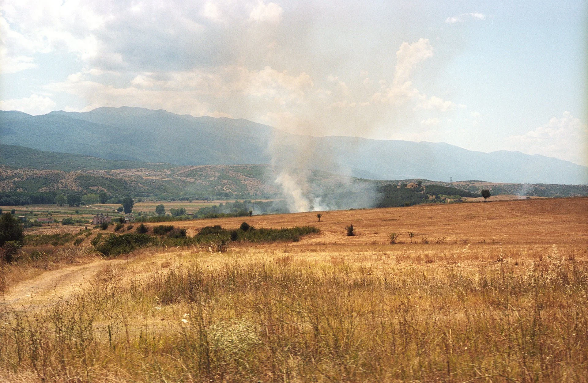 Open field with dry grass in the foreground, smoke rising from a fire in the middle ground, hills and mountains in the background under a partly cloudy sky.