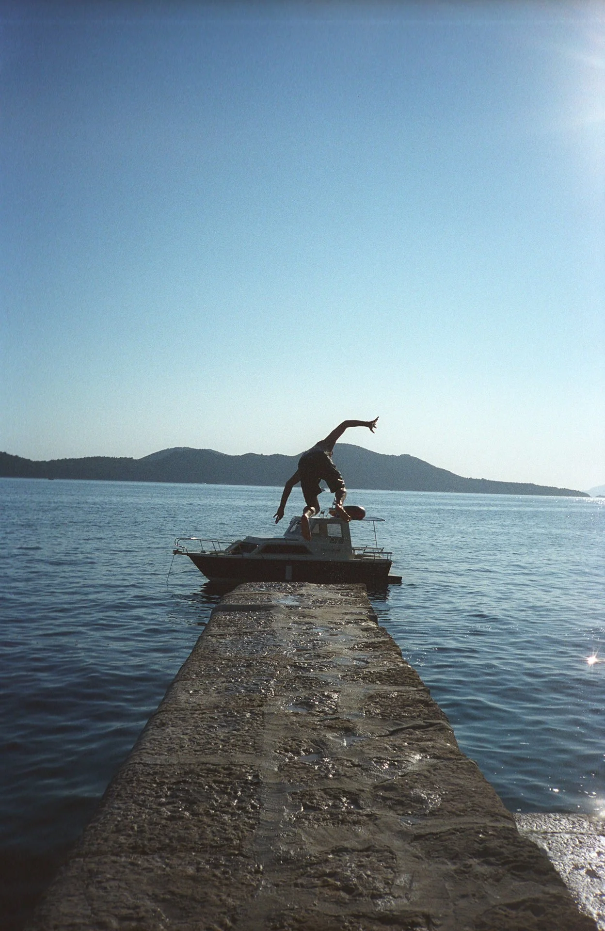 Person performing a handstand on a boat docked at a pier, with water and mountains in the background on a sunny day.