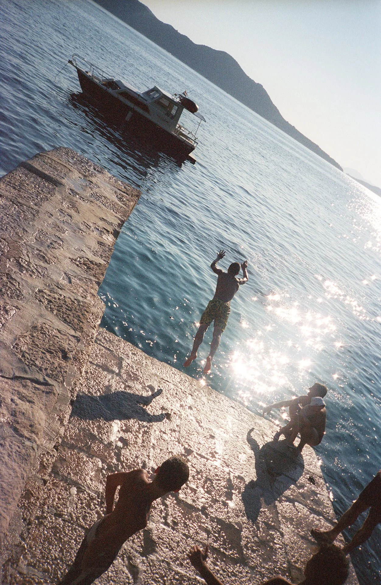 People swimming and playing in the water near a stone dock, with boats on the water and mountains in the background during sunset.