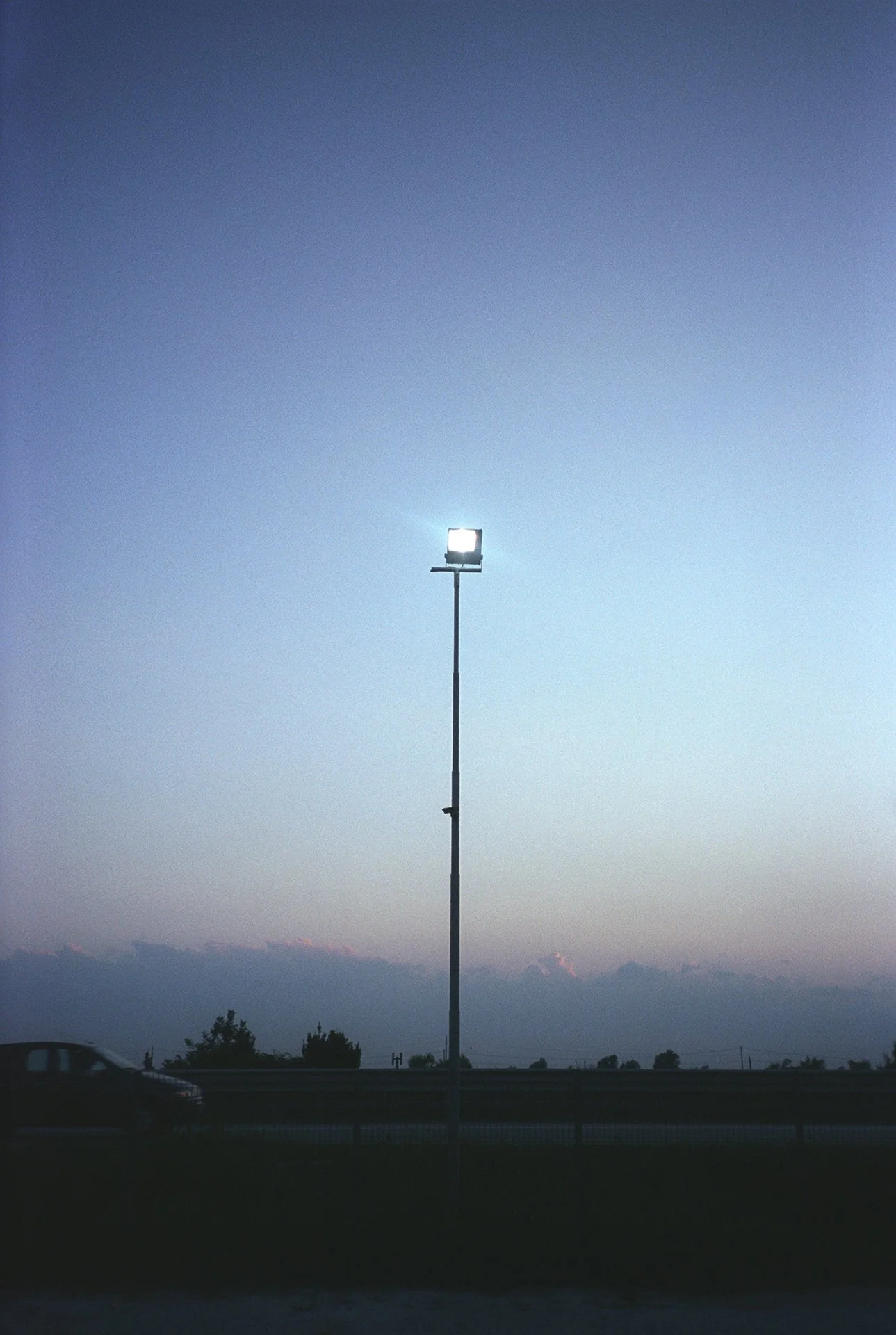 A tall stadium light pole with a single bright light against a dusk sky, with a car on a road and a distant landscape of hills or mountains.