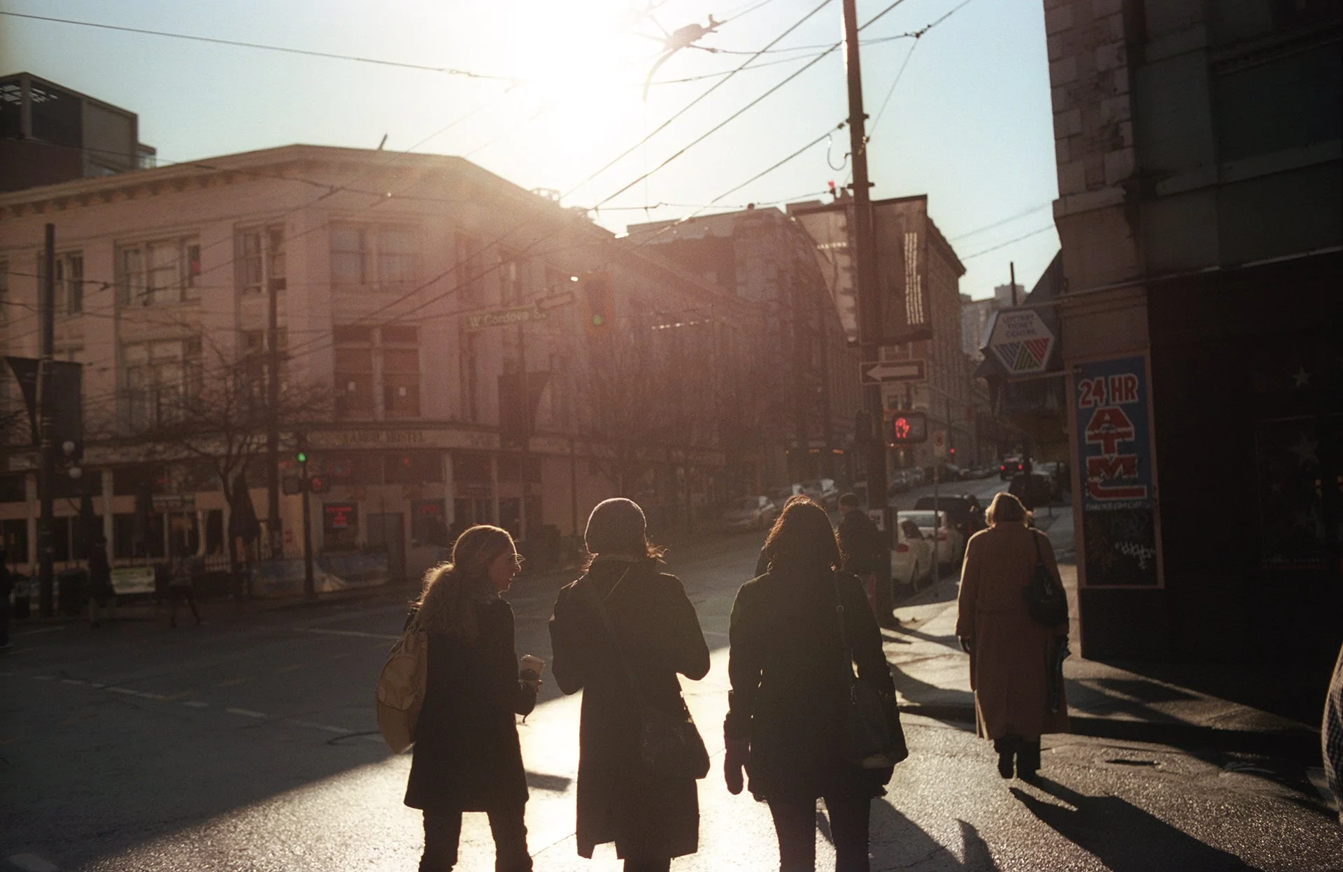 People crossing a city street at sunset, with buildings, traffic signals, and streetcar wires visible.