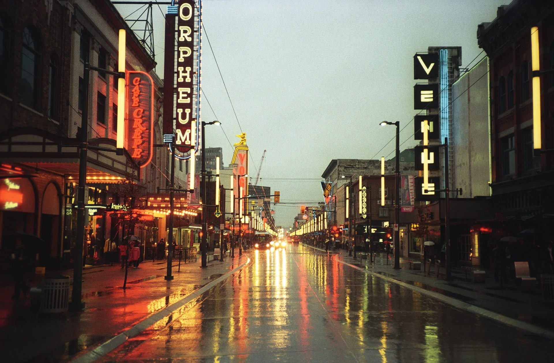 City street scene during rainy evening with neon signs on buildings, wet reflective pavement, and people walking with umbrellas.