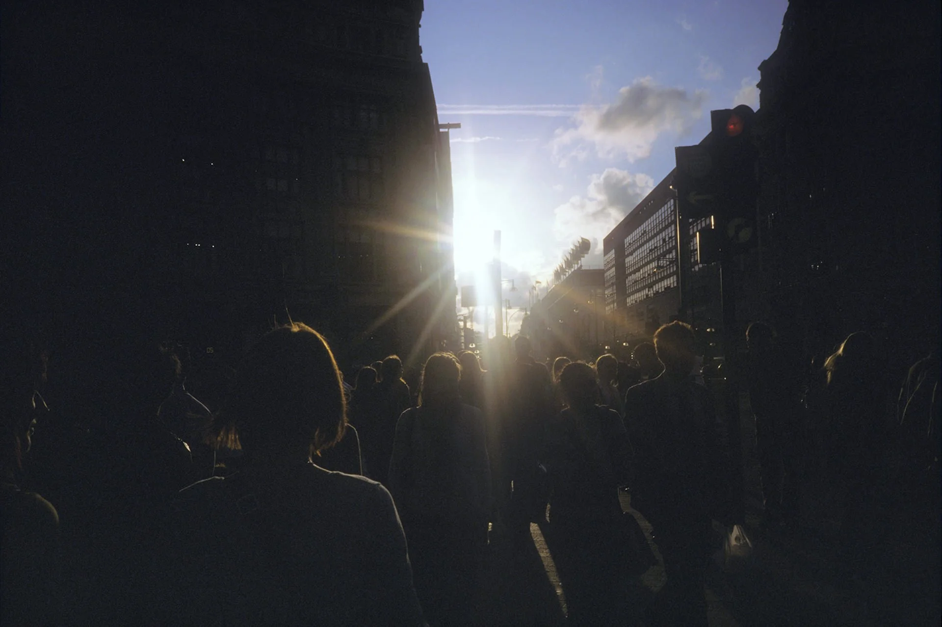 People standing on a city street at sunset, with dark buildings on either side and the sun shining brightly in the background.