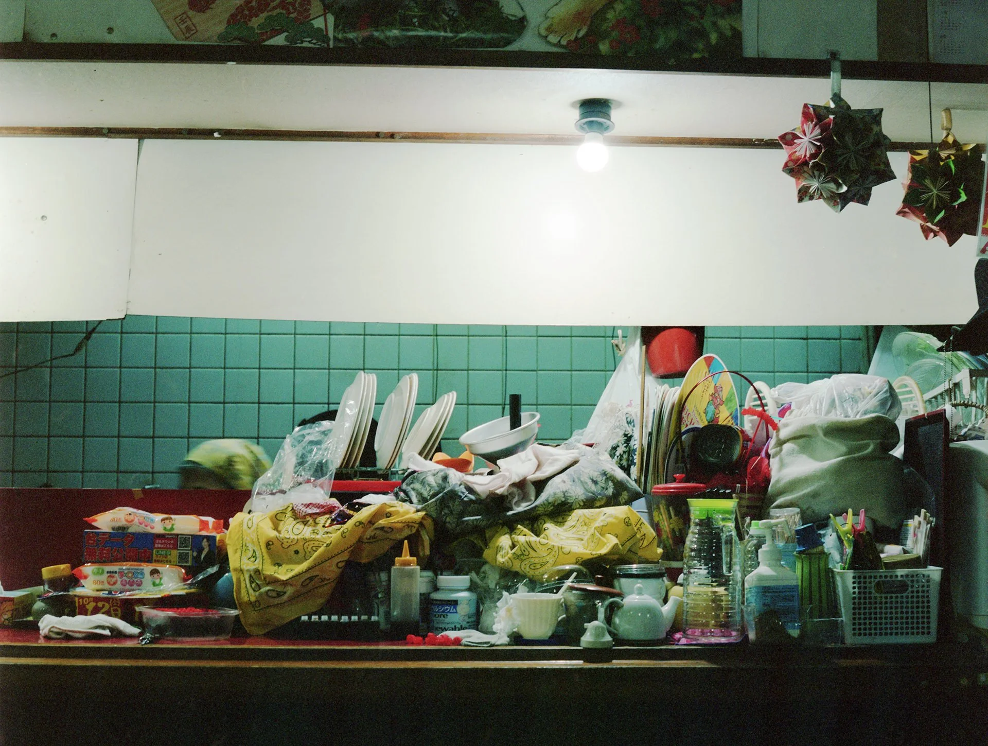 A cluttered kitchen counter filled with various objects including dishes, utensils, plastic bottles, cups, and food items. Overhead, a white cabinet and a ceiling light fixture are visible, with colorful decorations hanging from the cabinet.