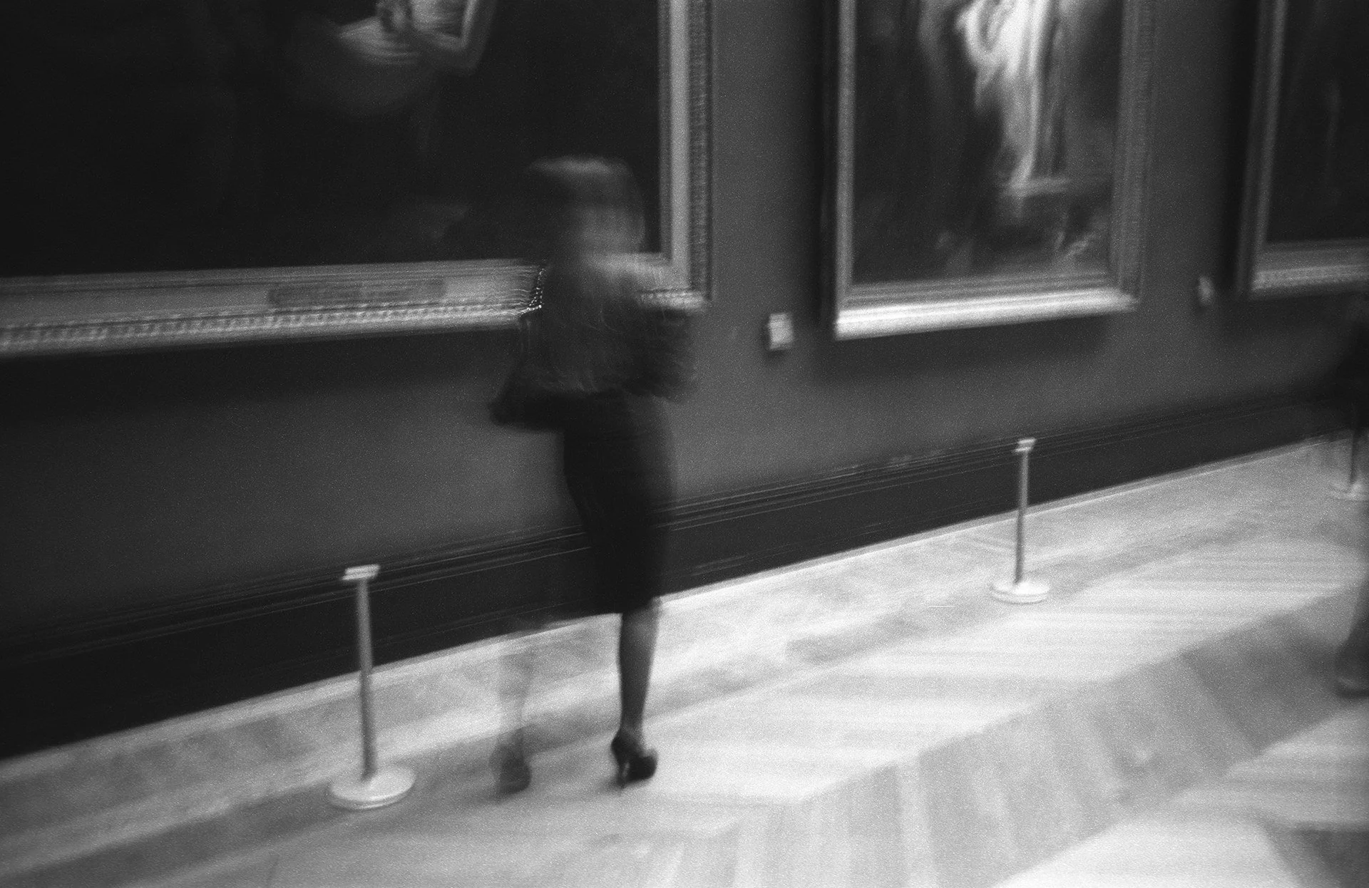 A woman dressed in historical clothing, possibly 19th or early 20th century, standing in front of framed paintings in a gallery or museum. The photo is black and white and slightly blurry.