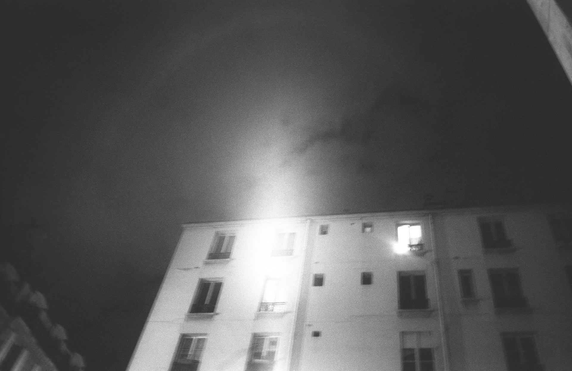 Nighttime view of a multi-story apartment building with some illuminated windows, and a bright light on the upper right side of the building, with a dark sky in the background.