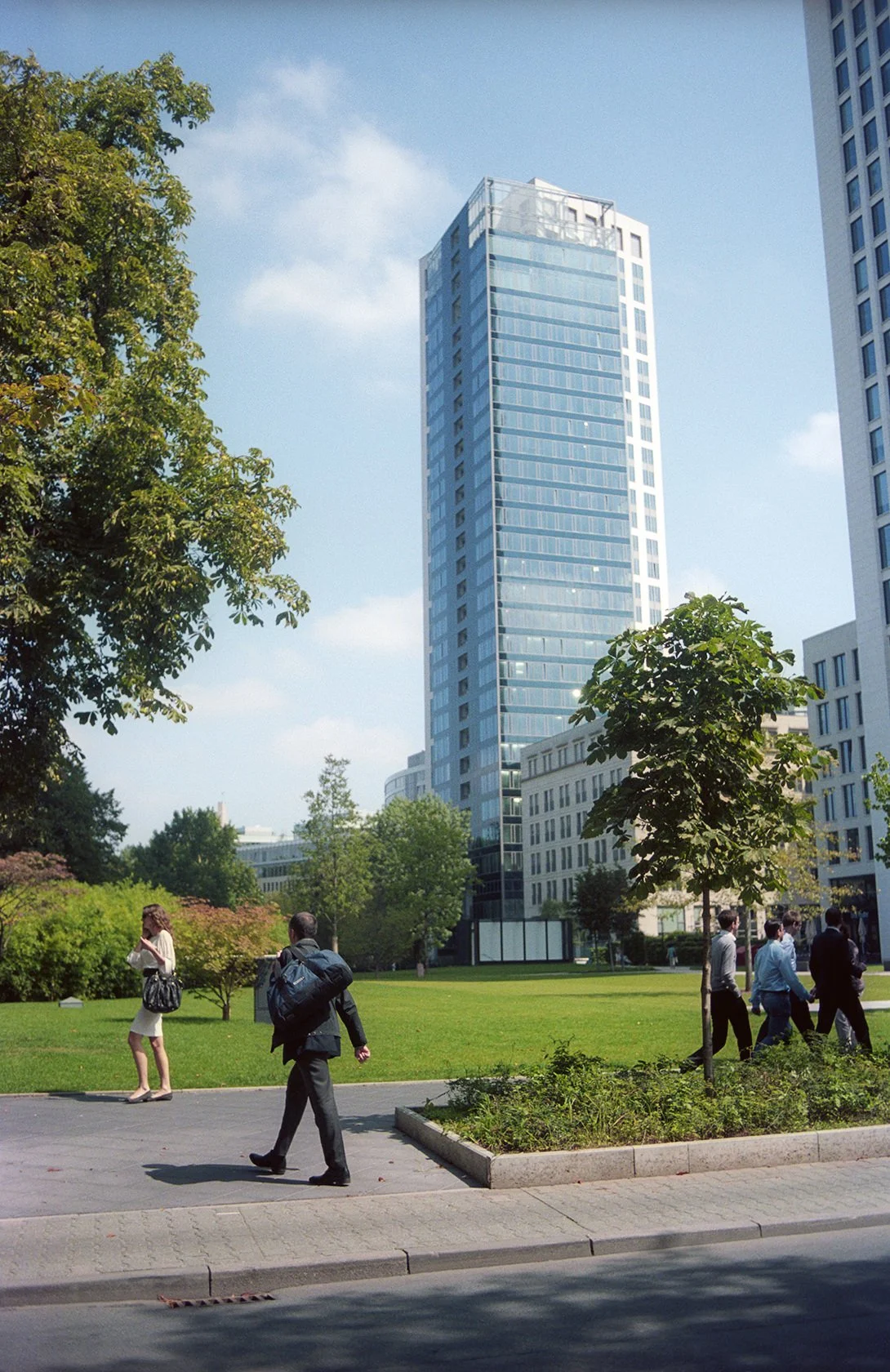 City park with tall modern glass building in the background, people walking and standing on the sidewalk, trees, and green grass.