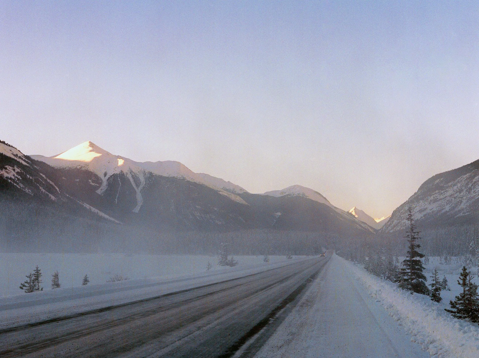 Snow-covered mountain range with a road in the foreground and a light blue sky.