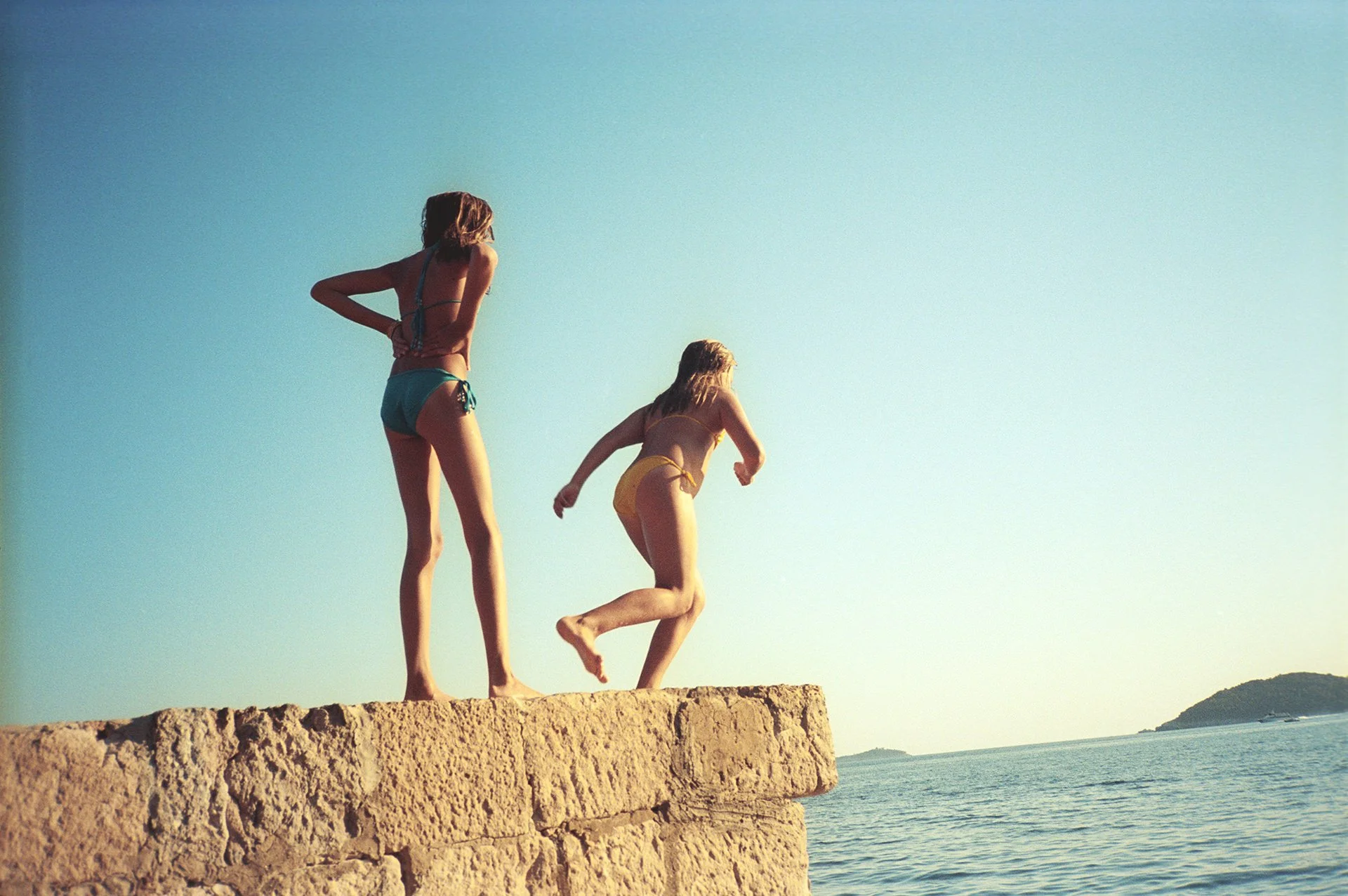 Two children in swimsuits jumping off a stone ledge into the water at a beach, with blue sky and ocean in the background.