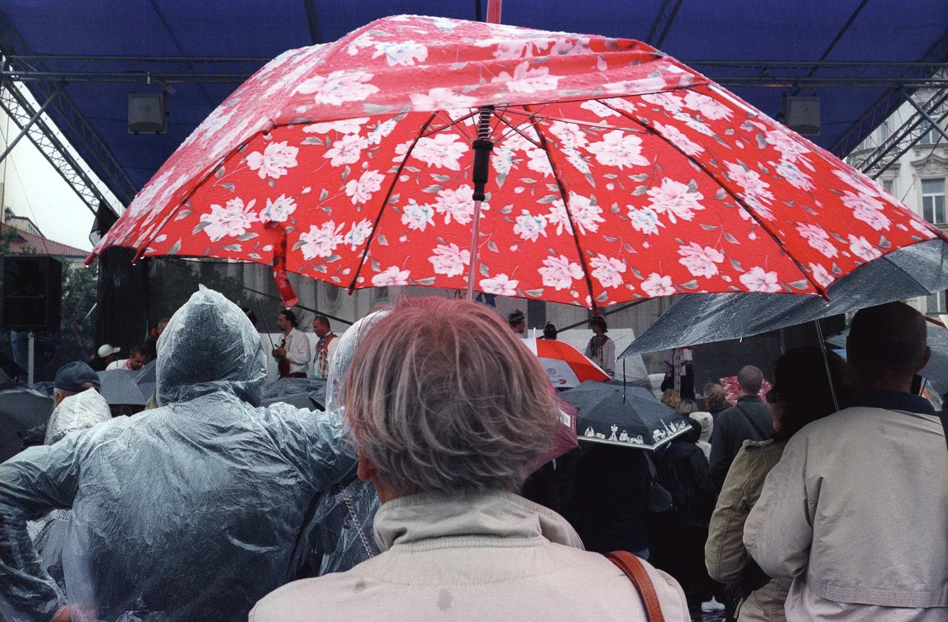 A crowd of people gathered under rain with some holding umbrellas, including a prominent bright red floral umbrella, at an outdoor event with a stage and building in the background.